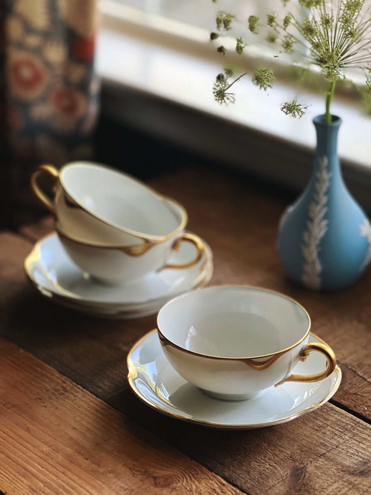 Antique teacups and saucers that are white with gold trim and a Wedgwood Jasperware vase is in the background