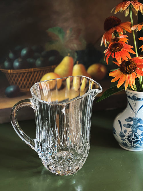 Clear glass pitcher on a table with fruits and flowers in the background