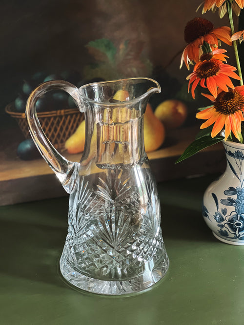 Clear glass pitcher on a table with flowers and fruits in the background