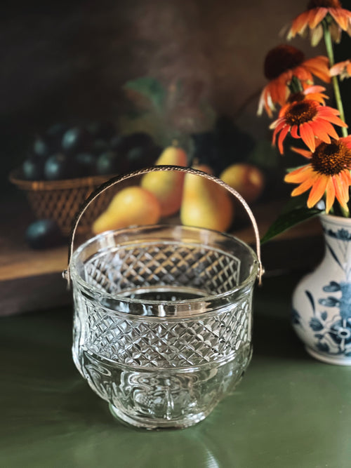 Clear glass basket with decorative handle on a table with fruits and flowers in the background
