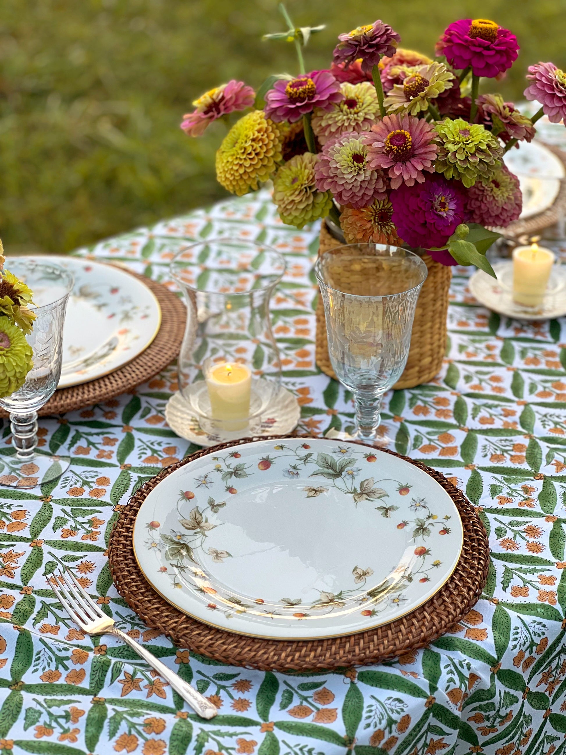 Outdoor table setting with a floral patterned tablecloth, white plates with gold and green botanical borders, woven brown placemats, clear glassware, and a wicker basket vase filled with pink, purple, and green flowers. A small candle sits on each plate.