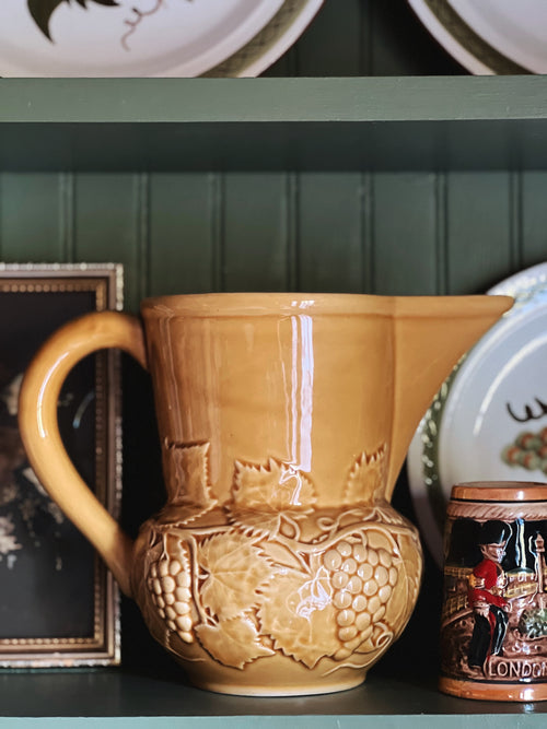 Ceramic pitcher with grapevine design on a shelf with decorative items.