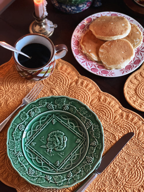 Table setting with green plates, a cup of coffee, and pancakes on a decorative tablecloth.