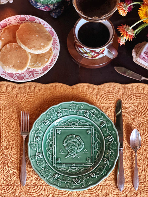Green decorative plate with a floral design on an orange textured tablecloth, accompanied by a cup of coffee and pancakes.