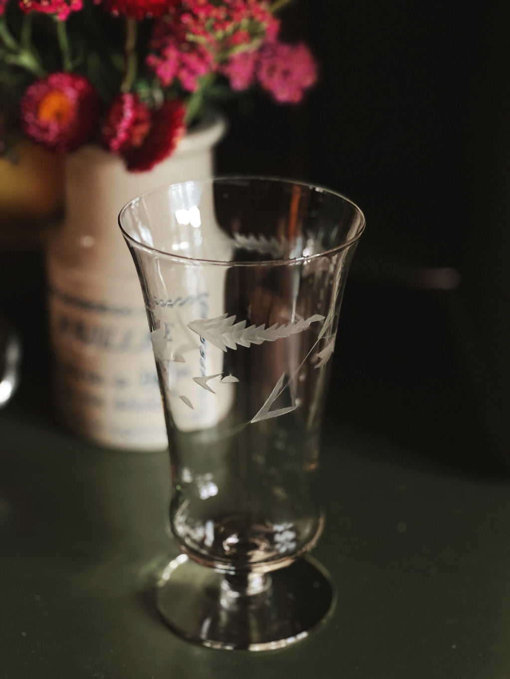 Decorative glass with engraved design on a dark surface, with flowers in the background.