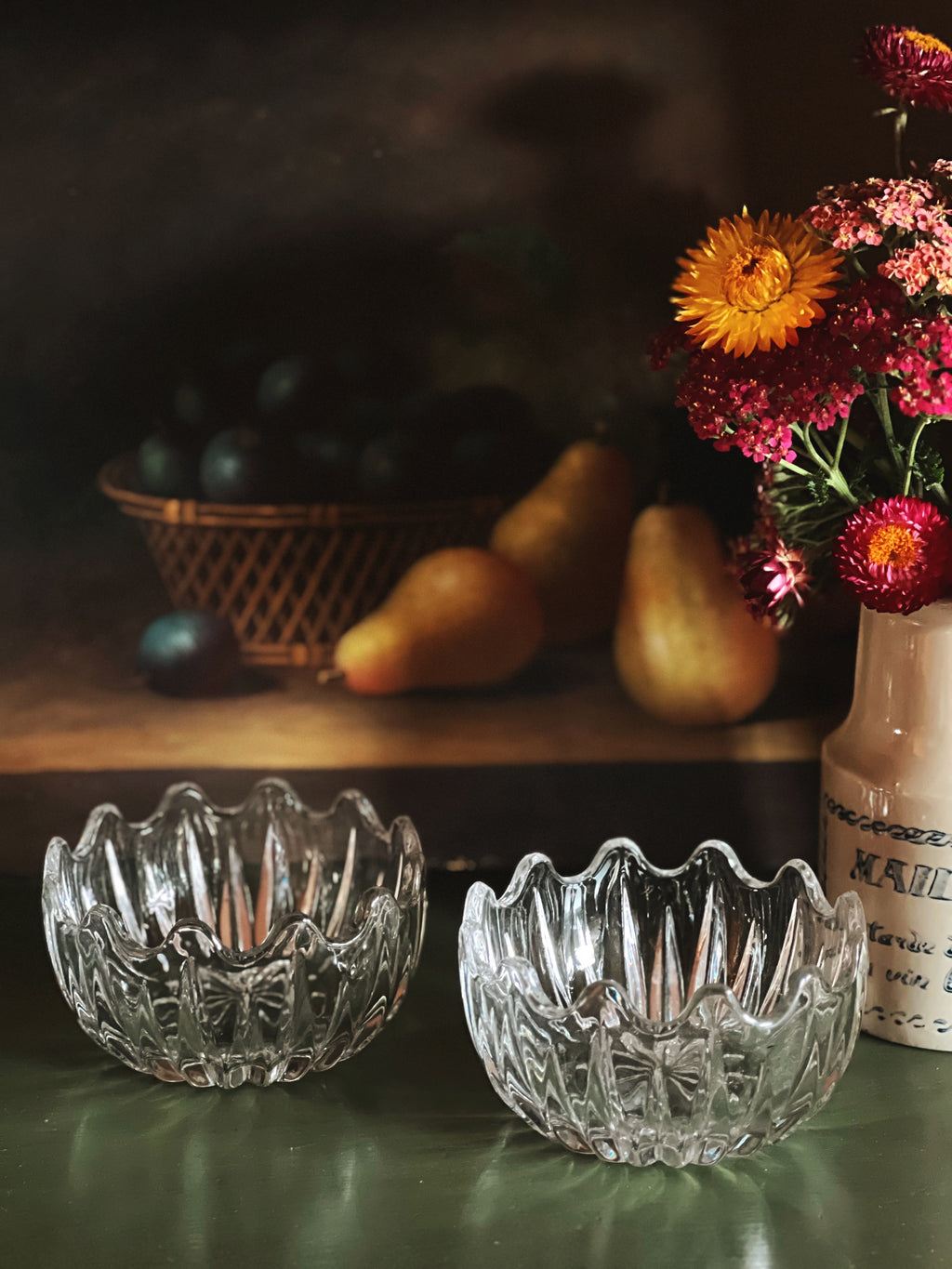 Two crystal bowls on a table with fruits and flowers in the background