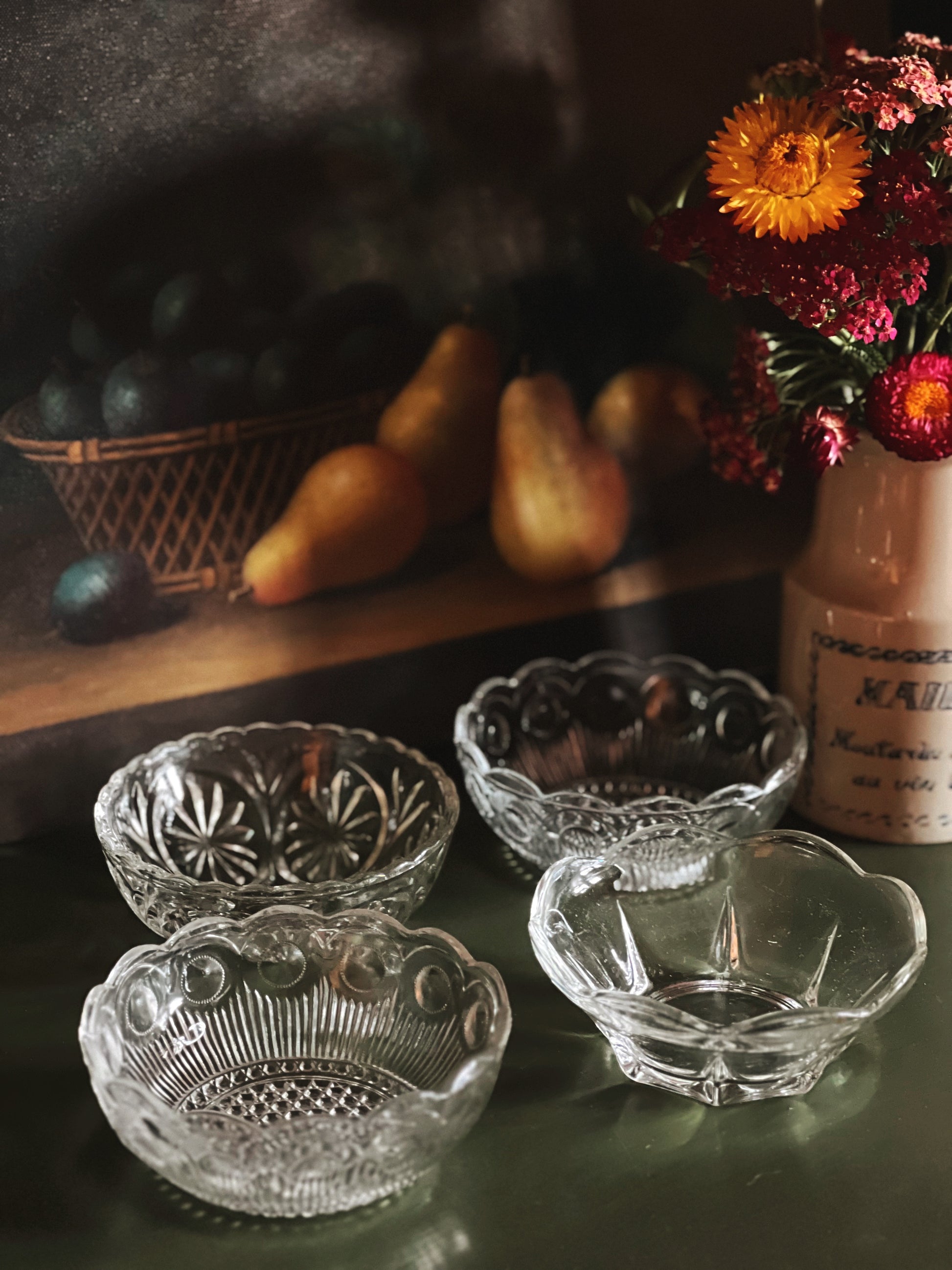 Set of four clear glass bowls on a dark surface with a blurred background featuring fruits and flowers.