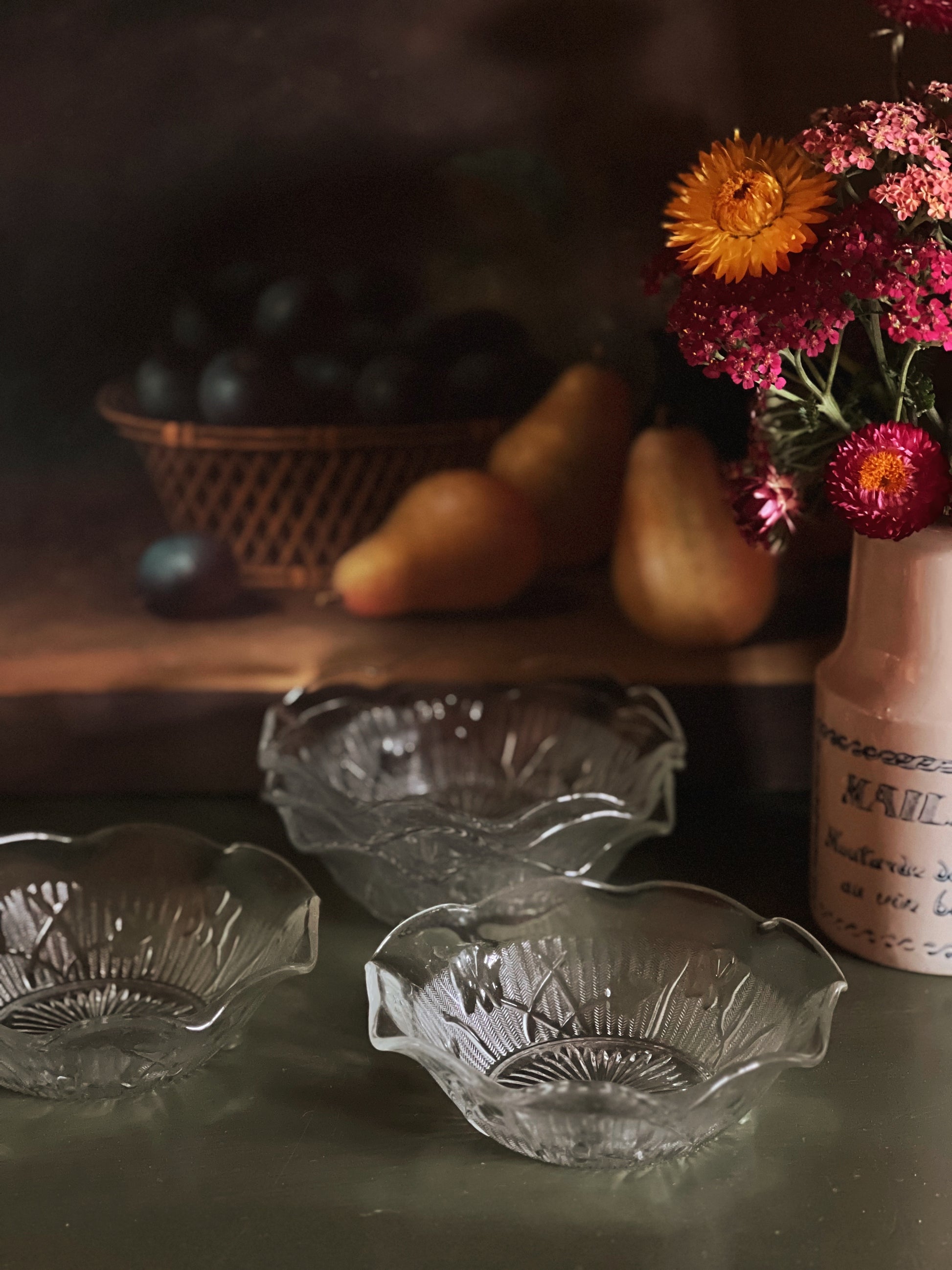 Clear glass bowls on a table with a vase of flowers and fruit in the background