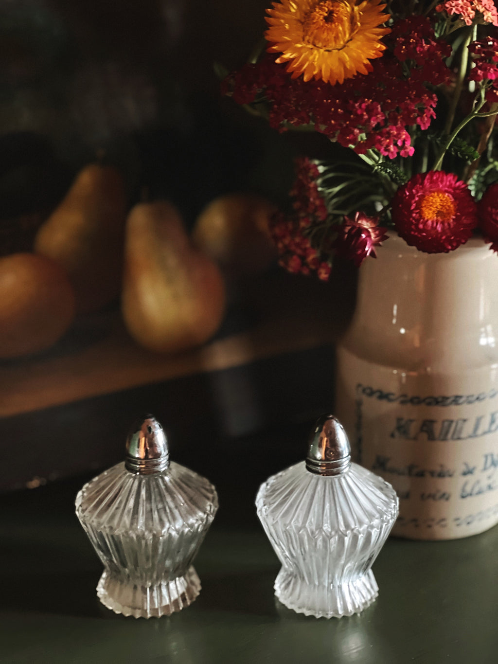 Two clear glass salt shakers with silver tops on a dark surface, with a vase of flowers and gourds in the background.