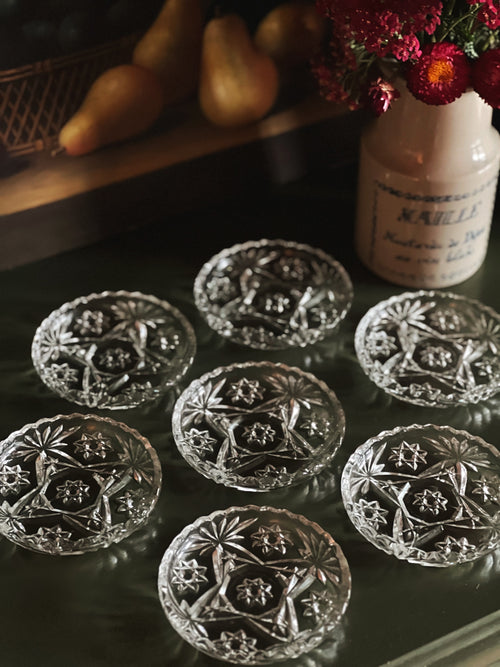 Set of crystal coasters with intricate designs on a dark surface, with a vase of flowers in the background.