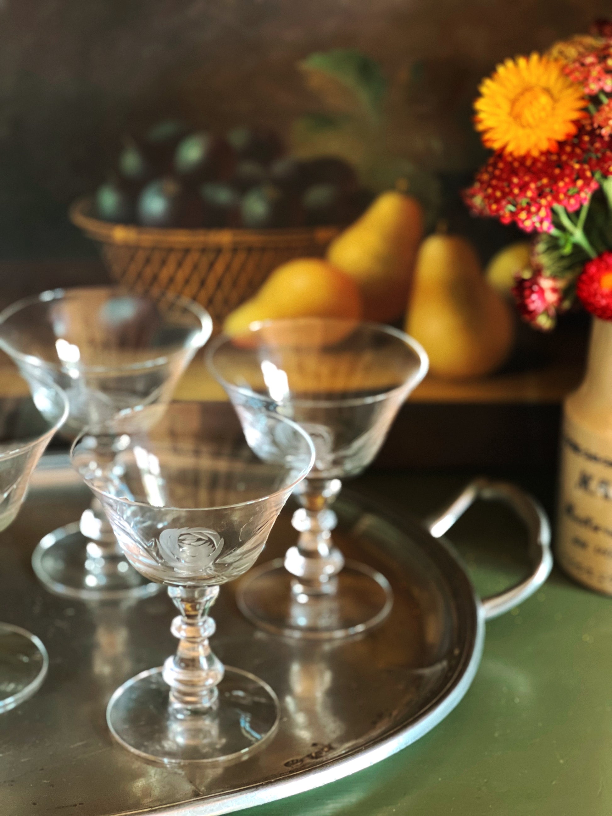 Clear glass goblets on a metal tray with fruits and flowers in the background