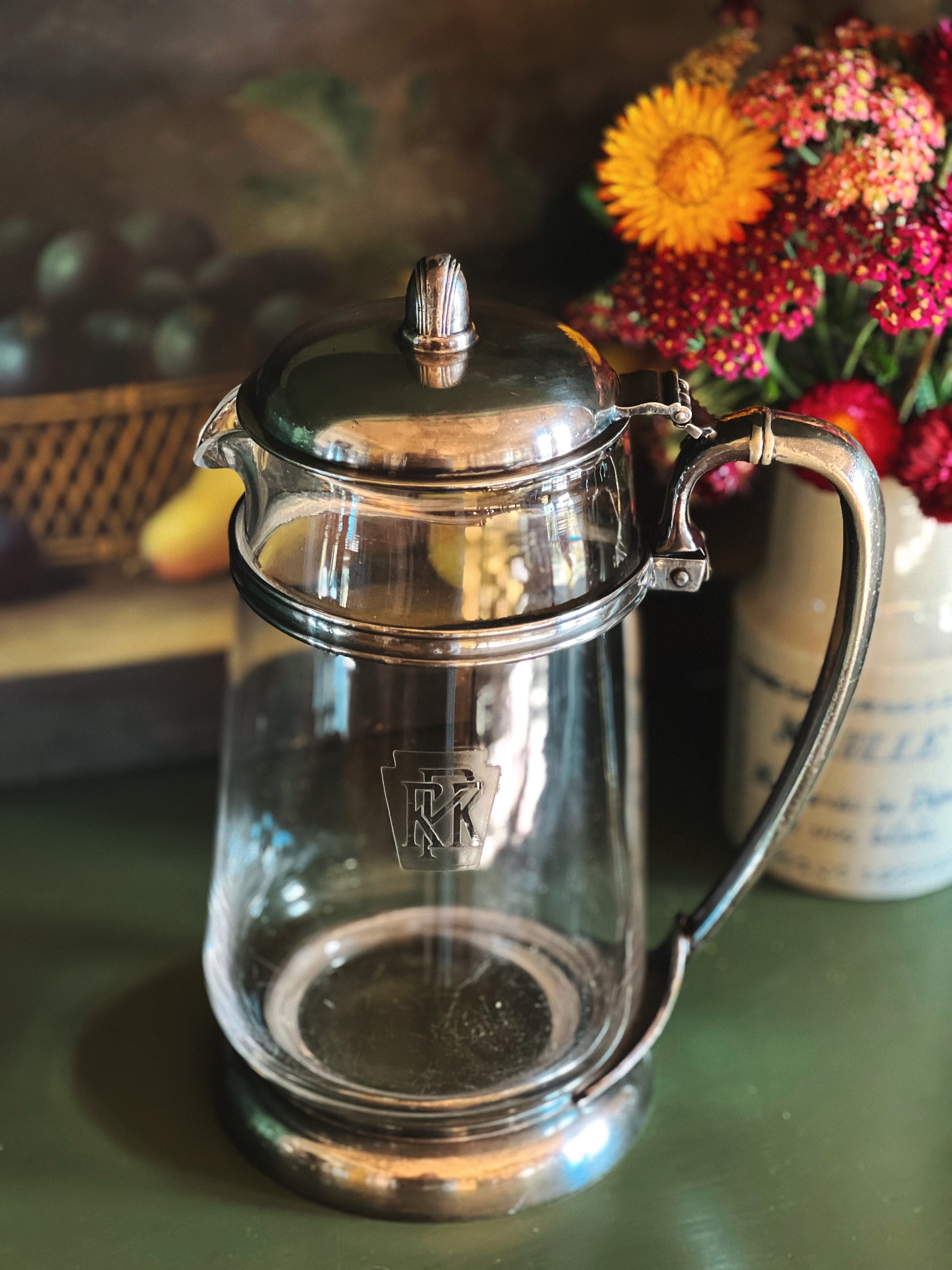 Clear glass teapot with silver lid and handle on a green surface with flowers in the background