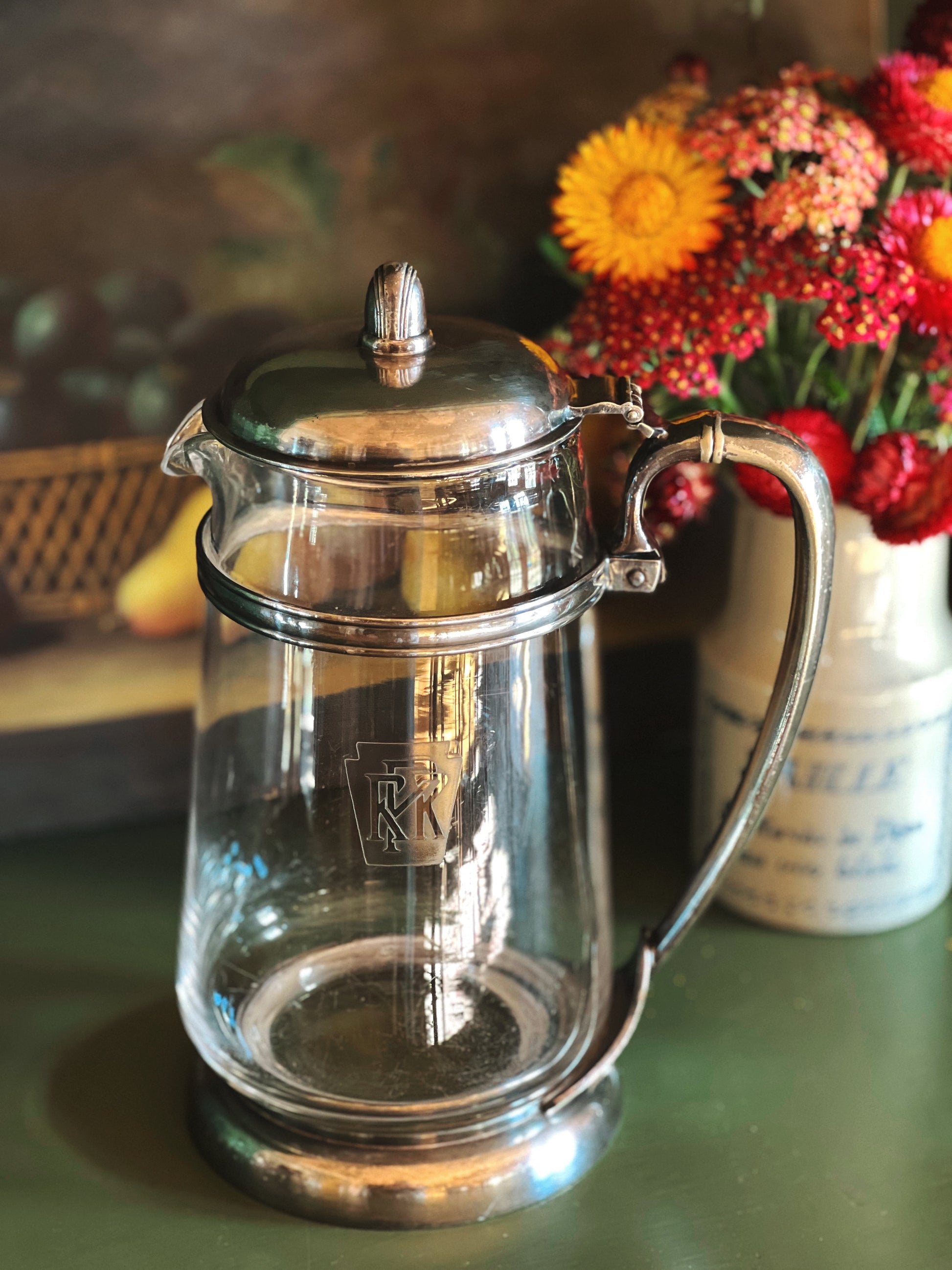 Clear glass teapot with silver lid and handle on a table with flowers in the background