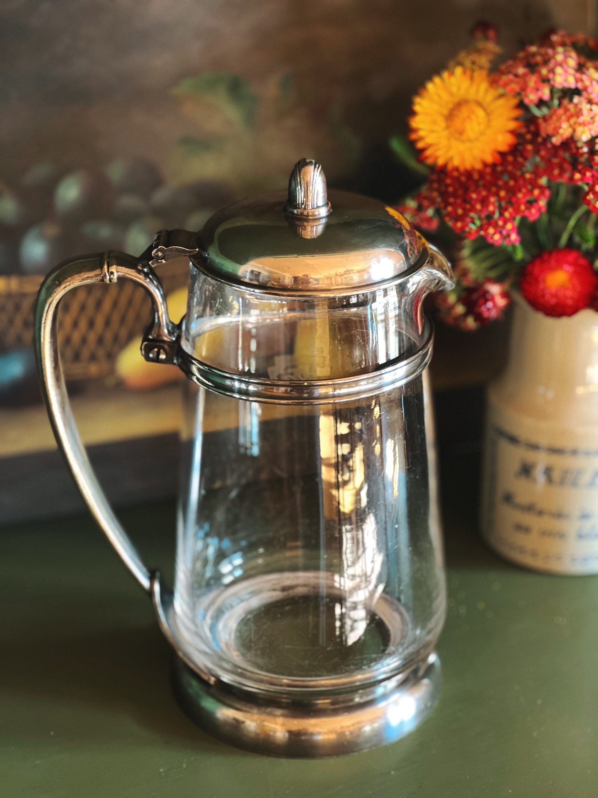 Clear glass teapot with silver lid and handle on a green surface with flowers in the background