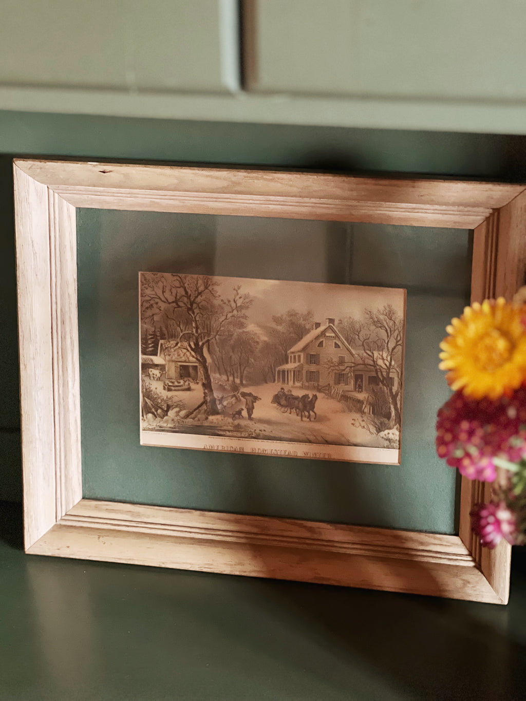Framed vintage-style landscape print on a green surface with flowers.