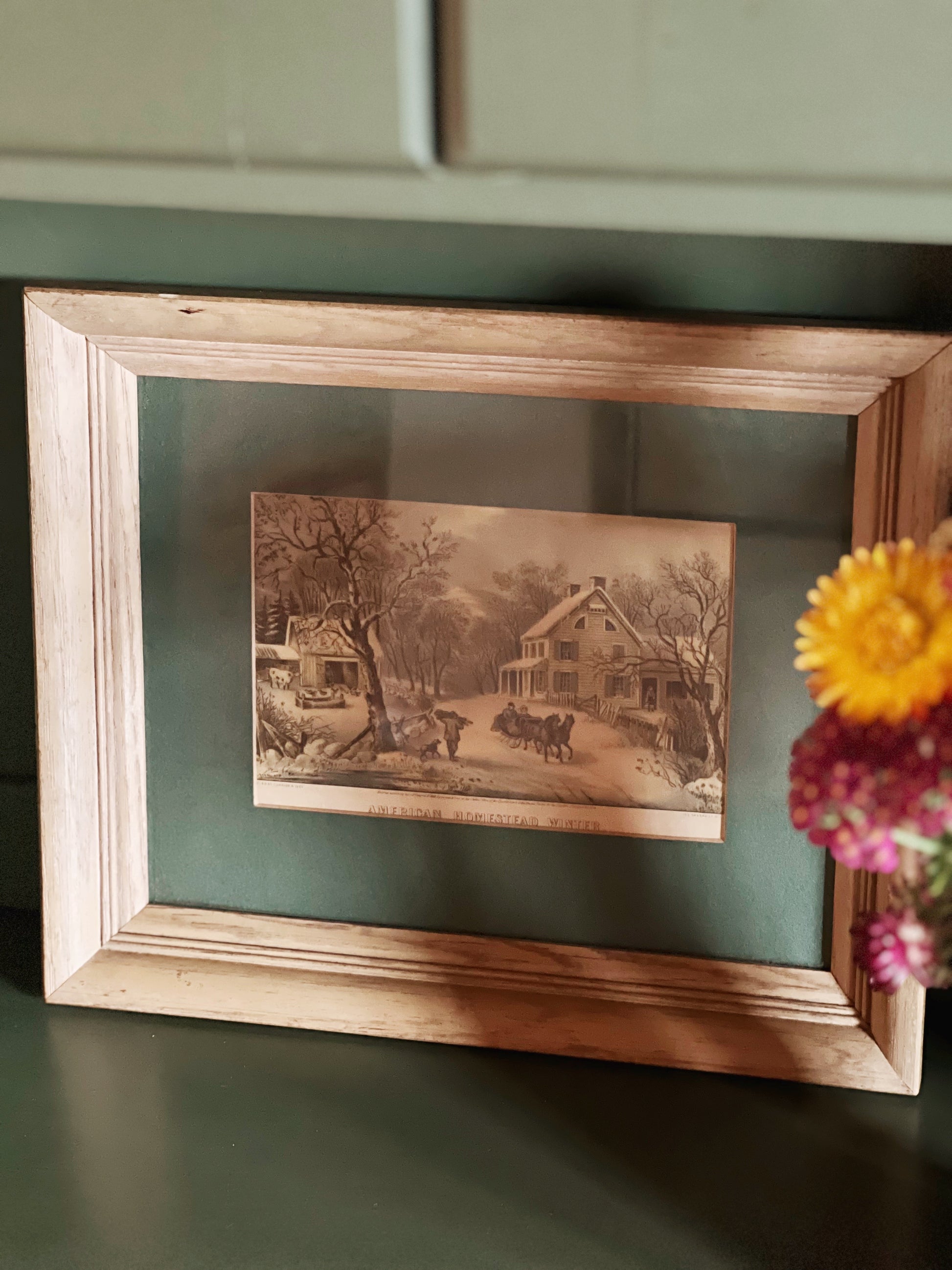 Framed vintage-style landscape print on a green surface with flowers.