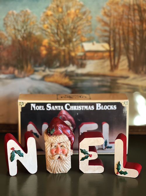 Wooden Christmas blocks spelling 'NOEL' with a Santa Claus face, in front of a decorative box labeled 'Noel Santa Christmas Blocks'.