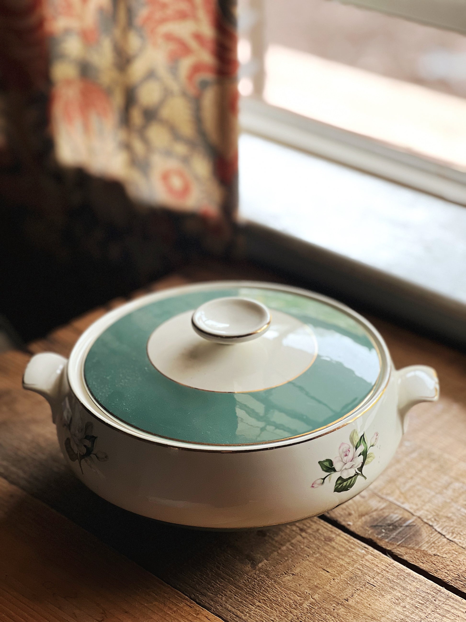 Ceramic pot with a green lid on a wooden surface near a window.
