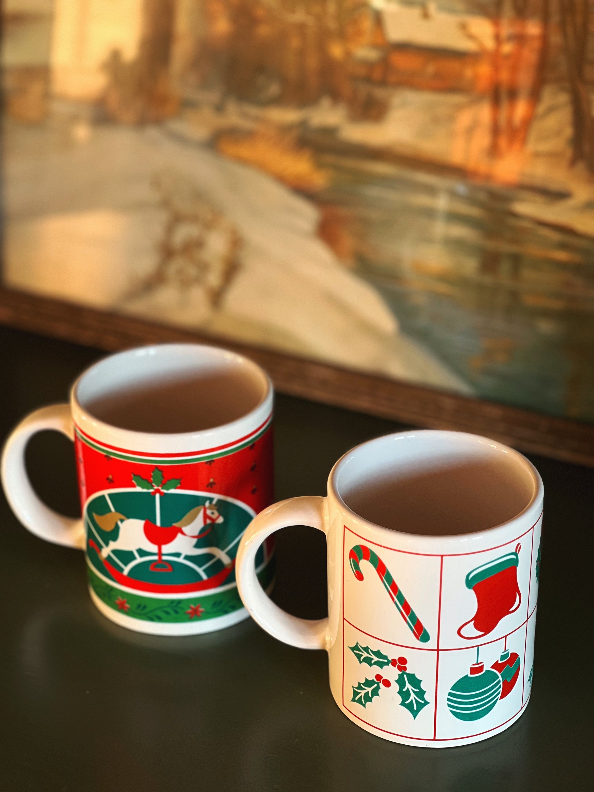 Two Christmas-themed mugs on a dark surface with a blurred landscape in the background.
