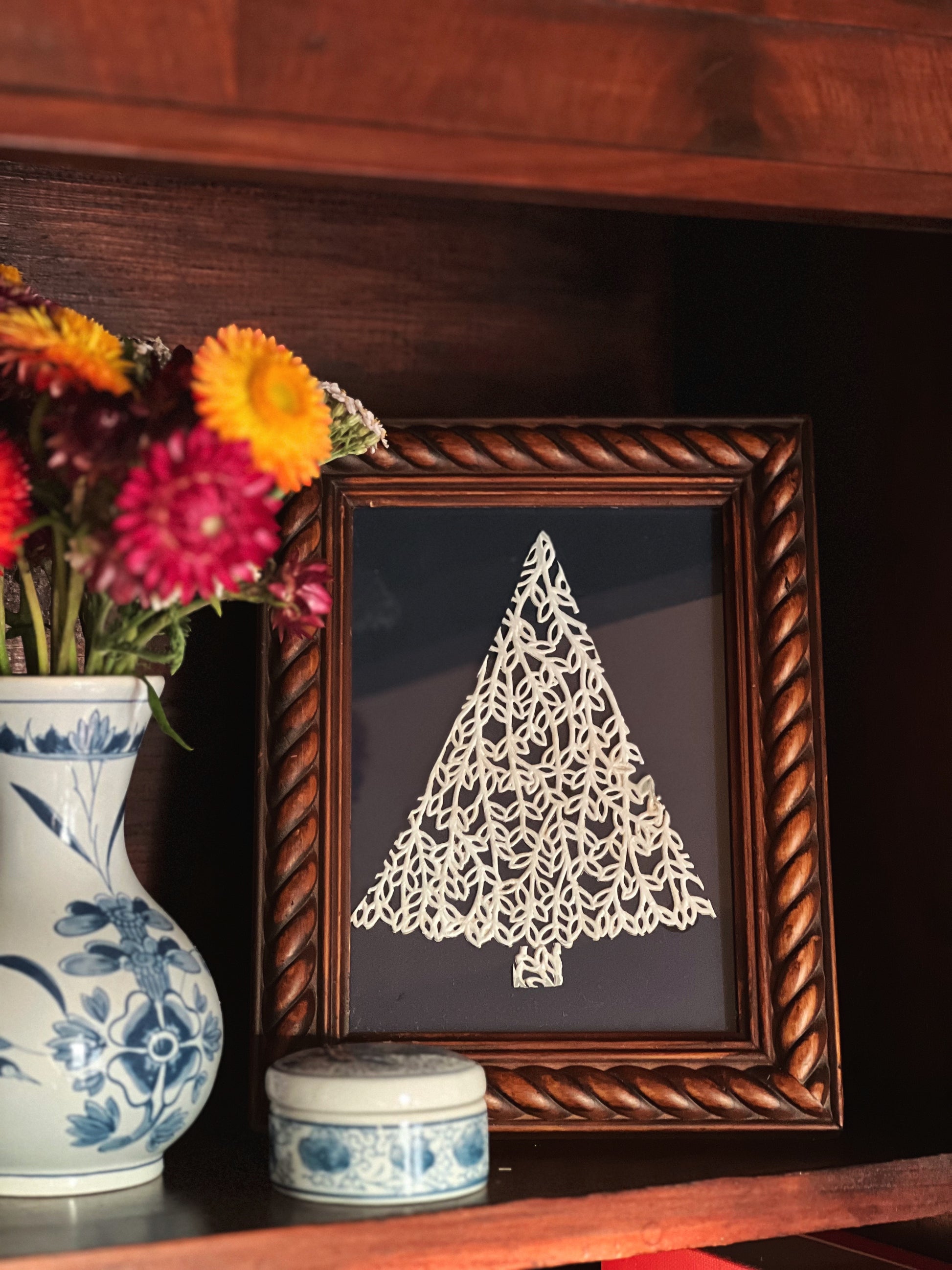 Framed paper tree decoration on a wooden surface with flowers and a vase.