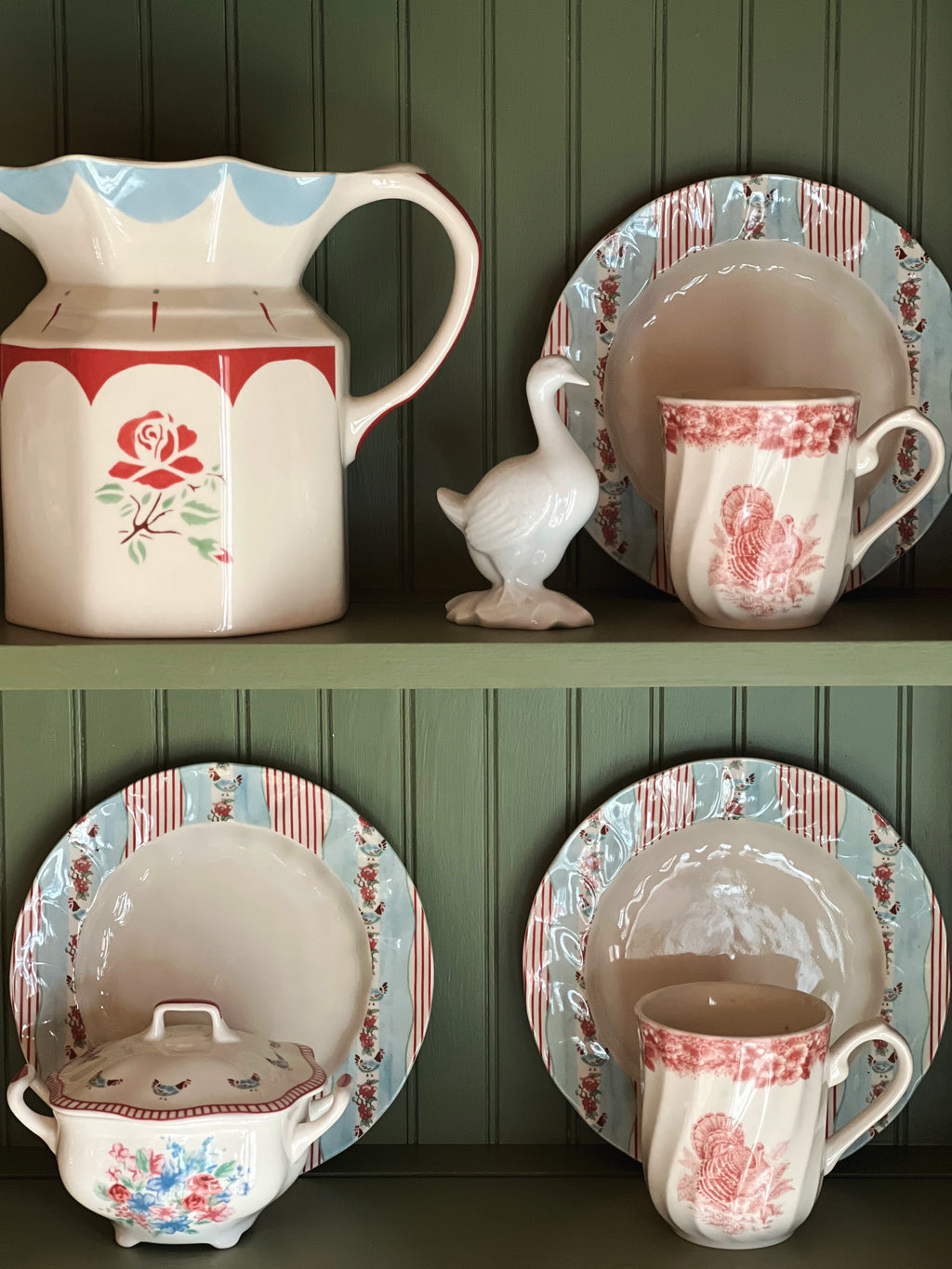 Collection of vintage-style ceramic dishes and a pitcher on a wooden shelf against a green paneled wall.