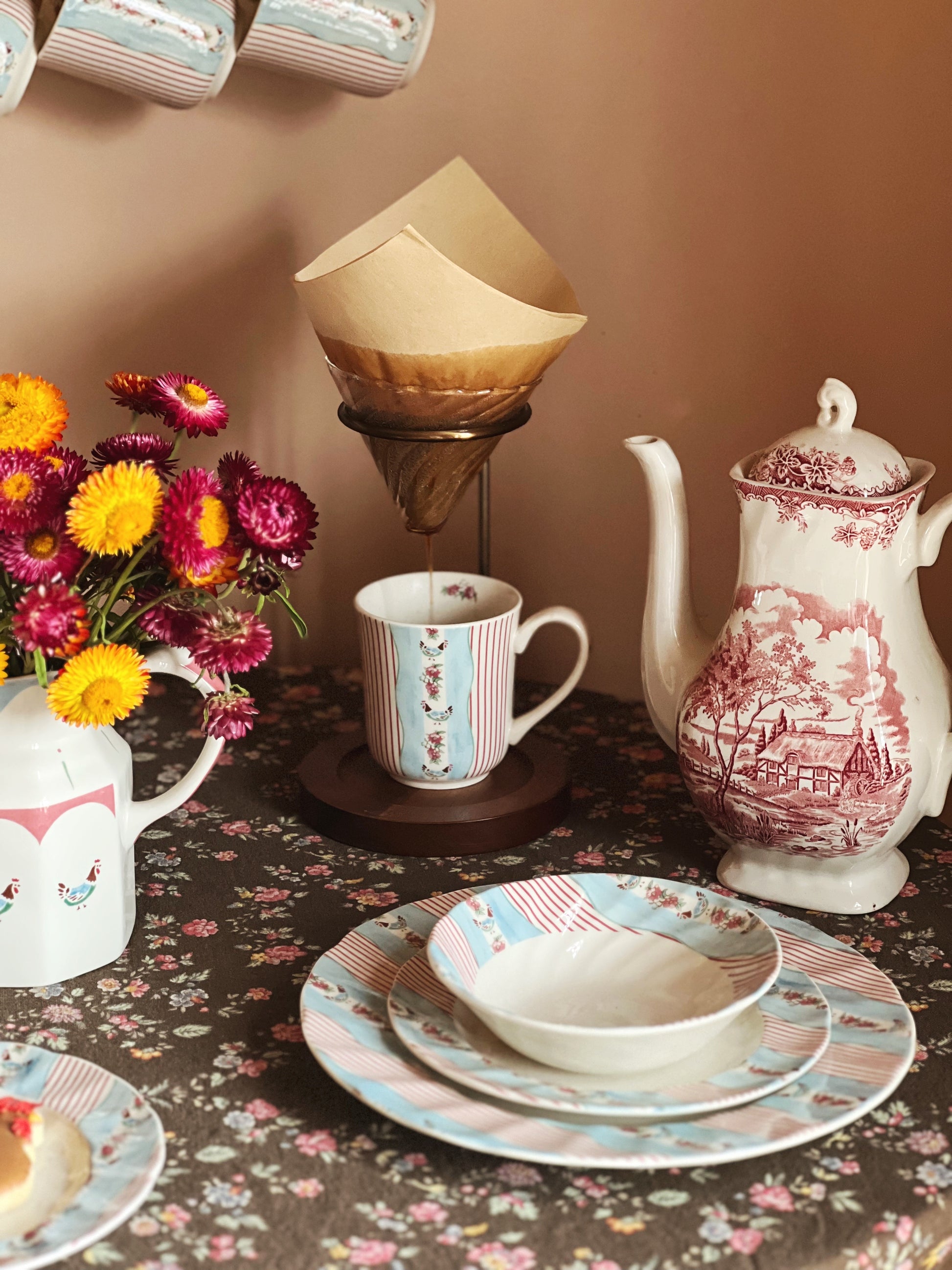 Tea set with teapot, cups, and saucers on a floral tablecloth with flowers and a candle in the background.
