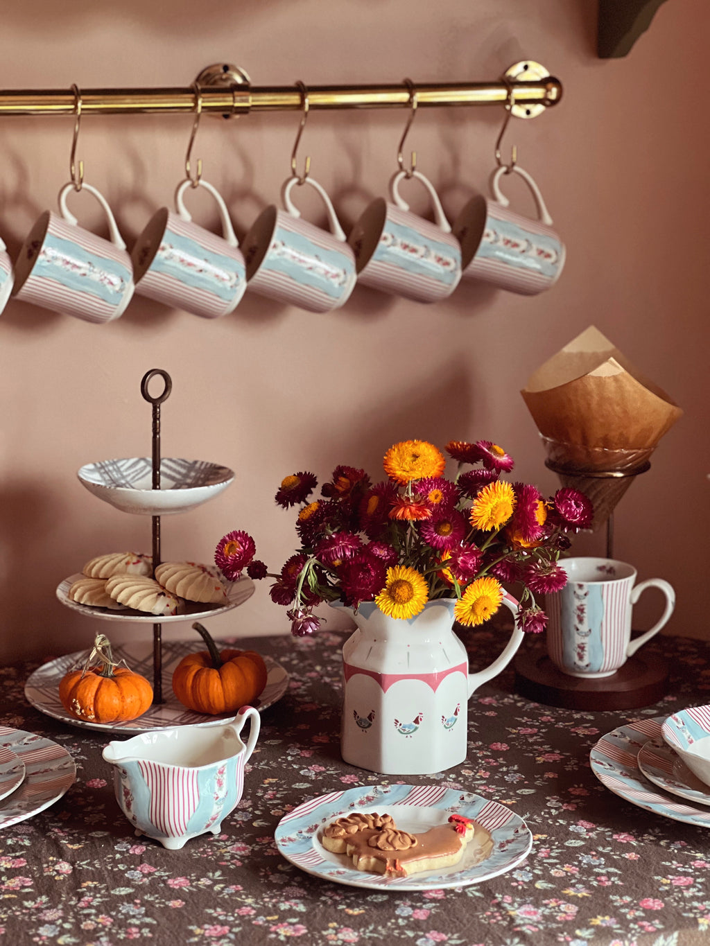Tea set with mugs, cookies, and flowers on a table against a wall with hanging mugs.
