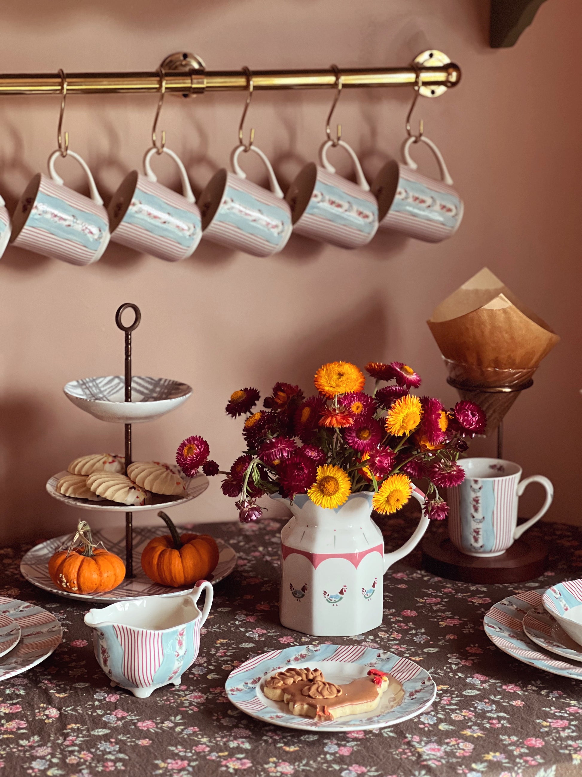 Tea set with mugs, cookies, and flowers on a table against a wall with hanging mugs.