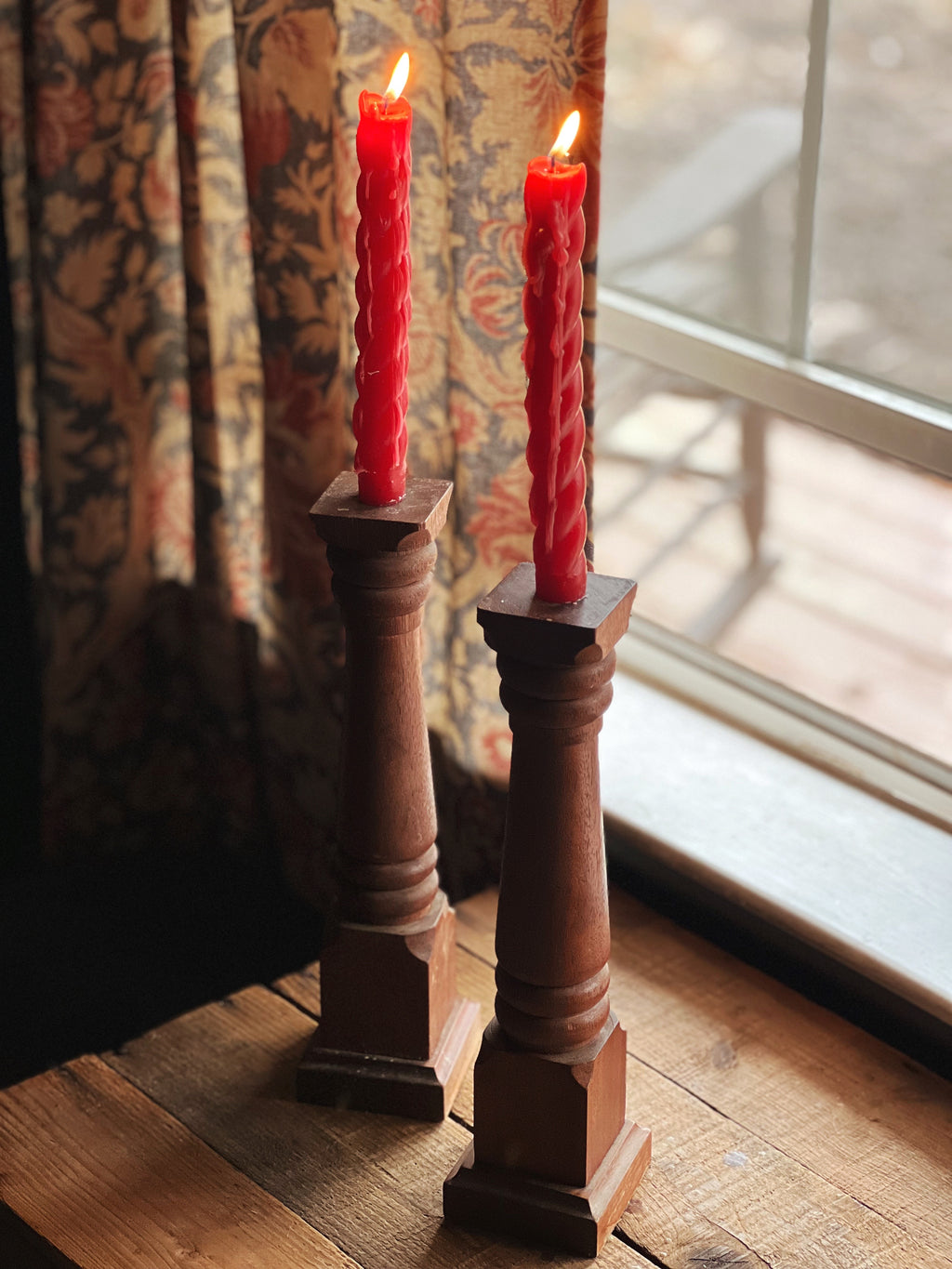 Two red candles in wooden candlesticks on a wooden floor with a patterned curtain and window in the background.