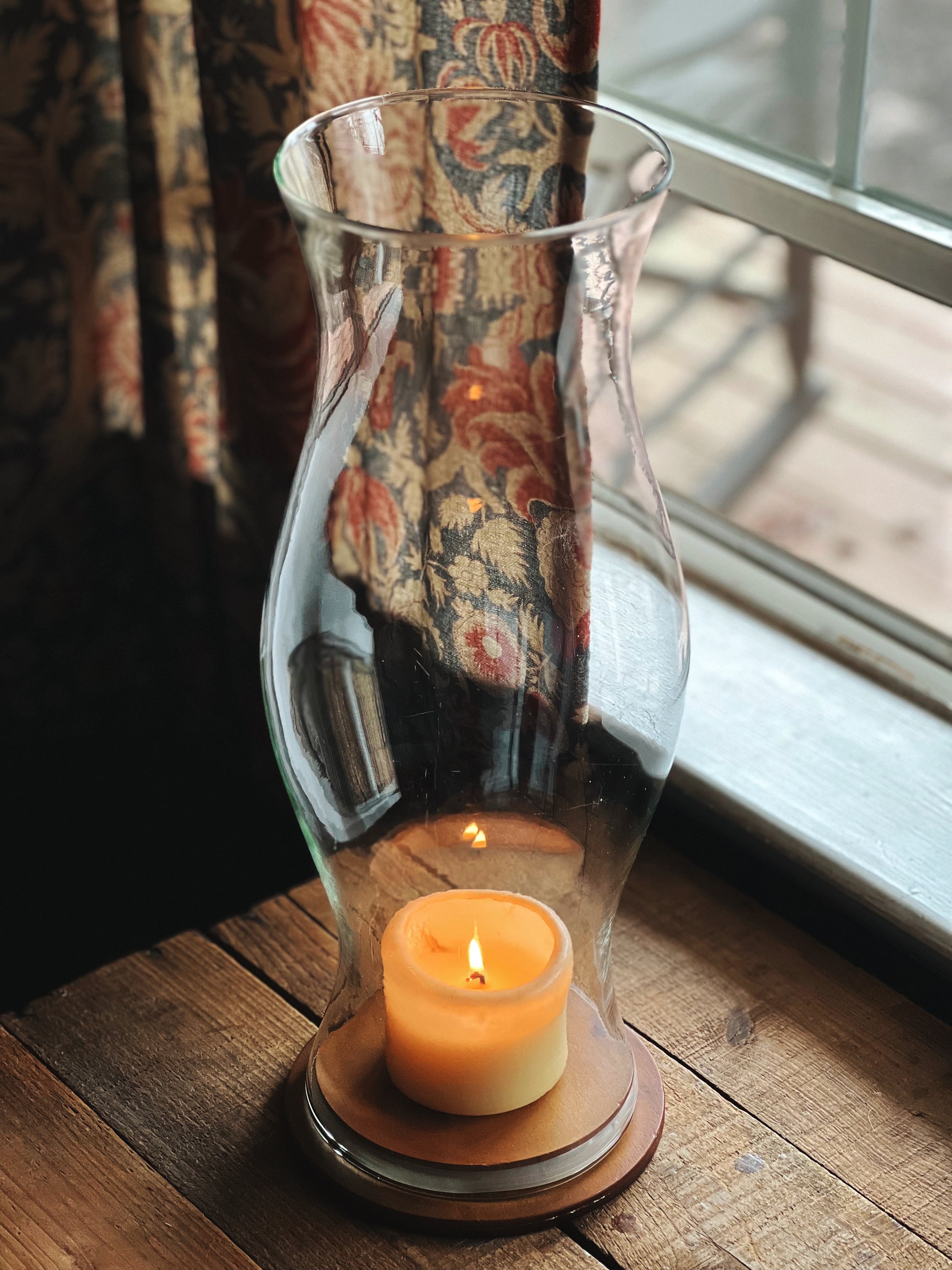 Candle in a glass lantern on a wooden surface with a floral curtain in the background