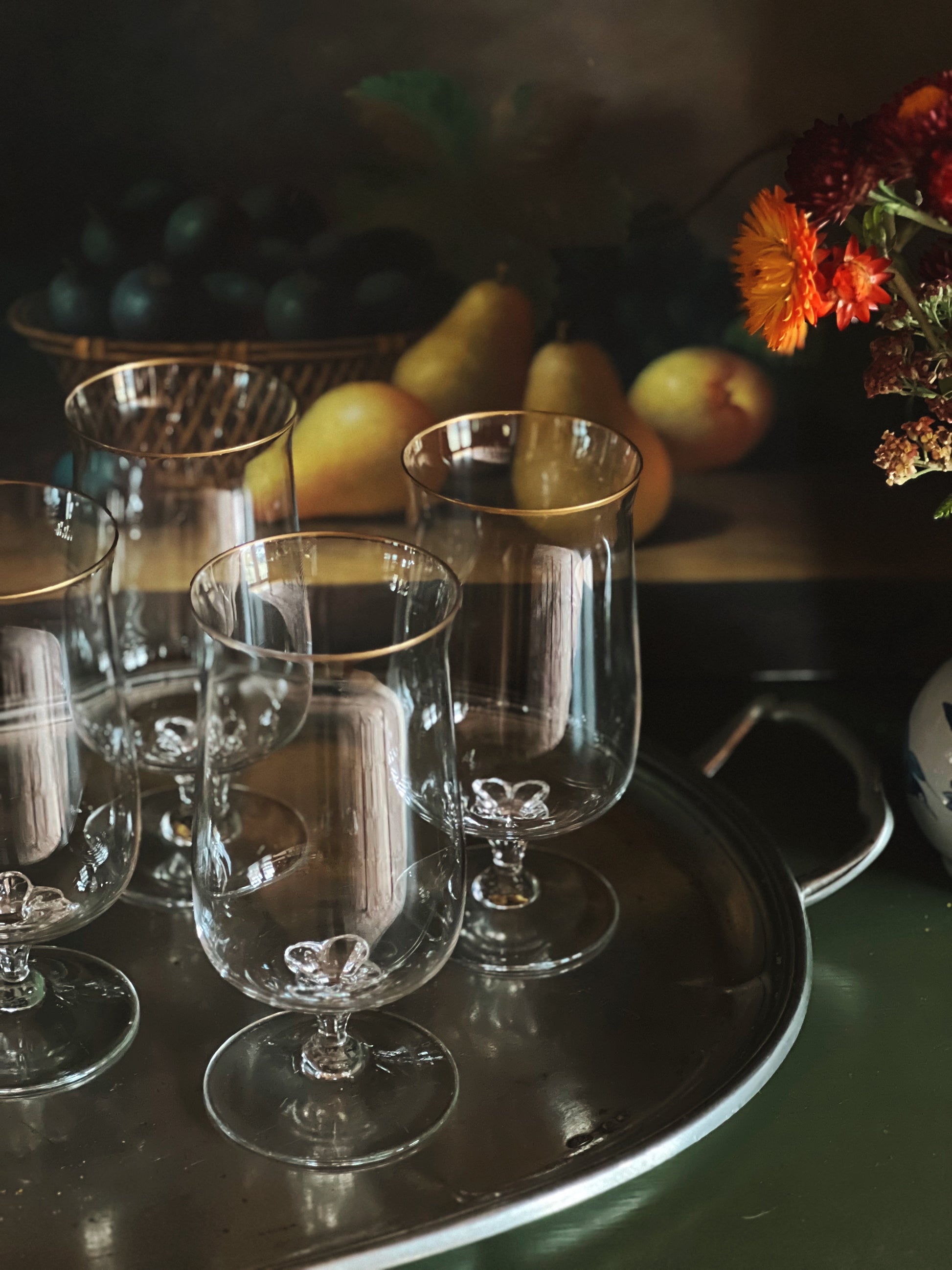 Set of clear glass goblets on a dark surface with fruits and flowers in the background