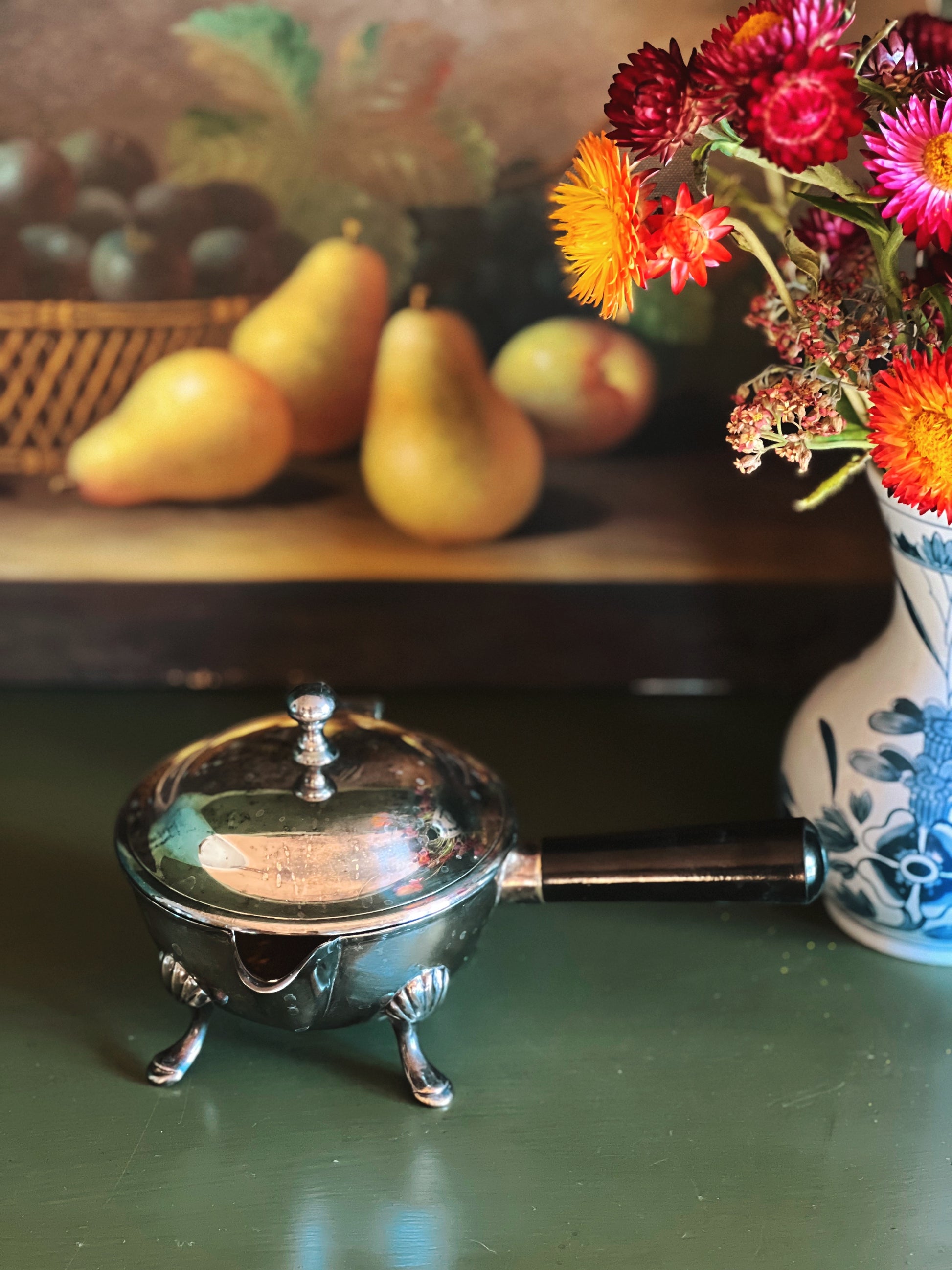 Silver container with a handle on a green surface, with pears and flowers in the background.