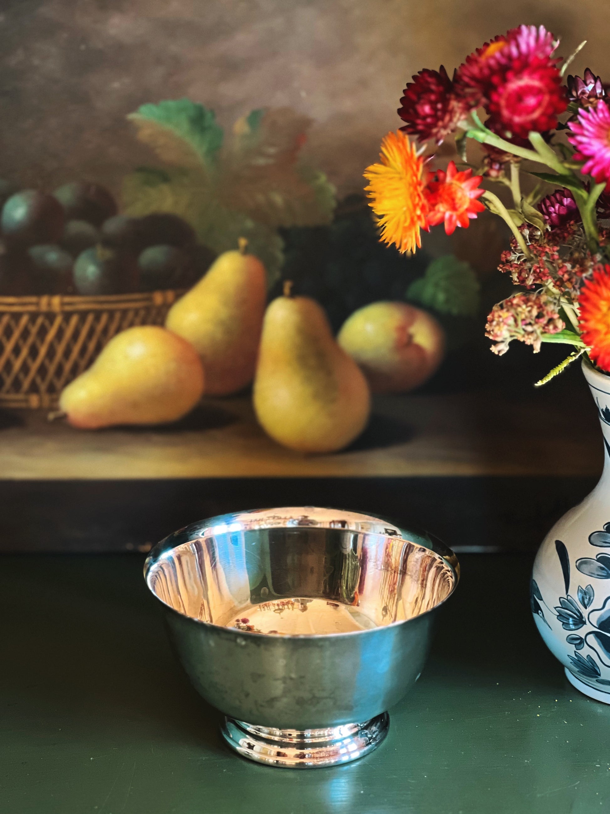 Silver bowl on a table with pears, grapes, and flowers in the background