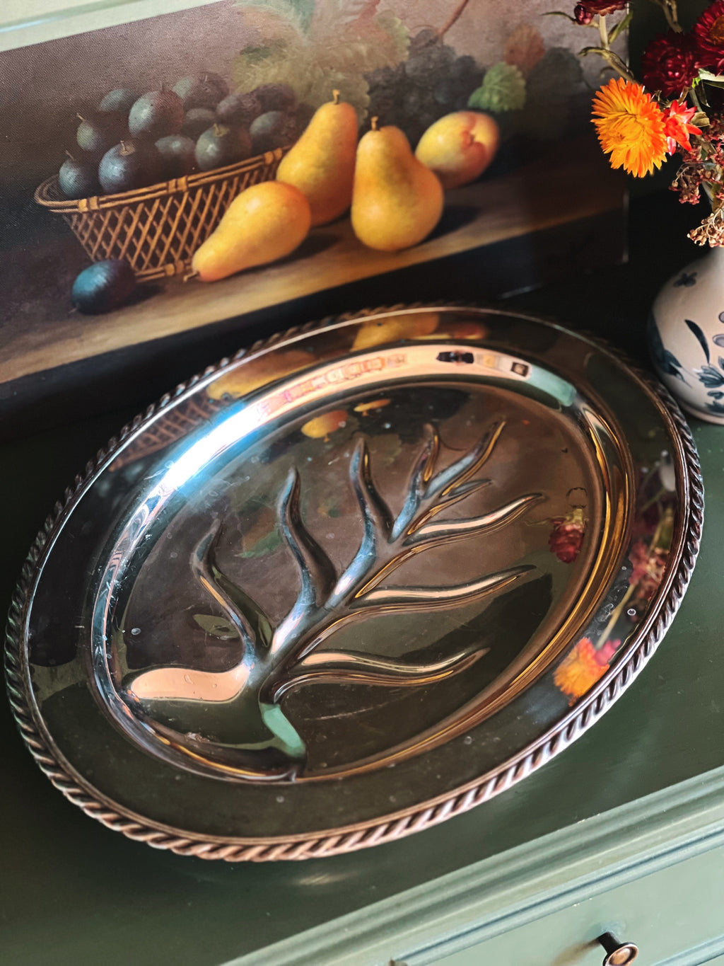 Decorative silver platter with tree design on a table with fruit and flowers in the background