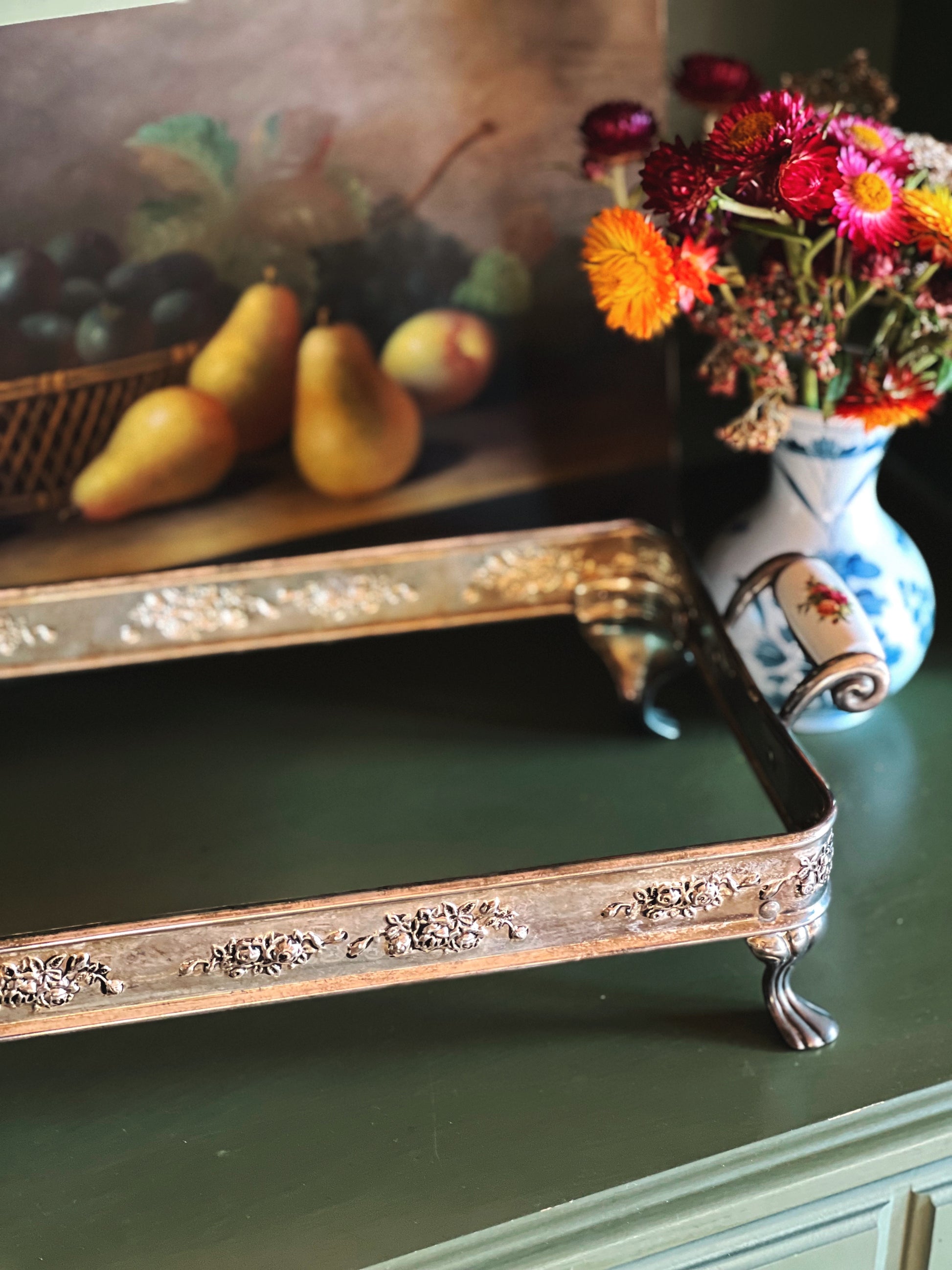 Decorative tray on a surface with a painting of fruit and a vase of flowers in the background.