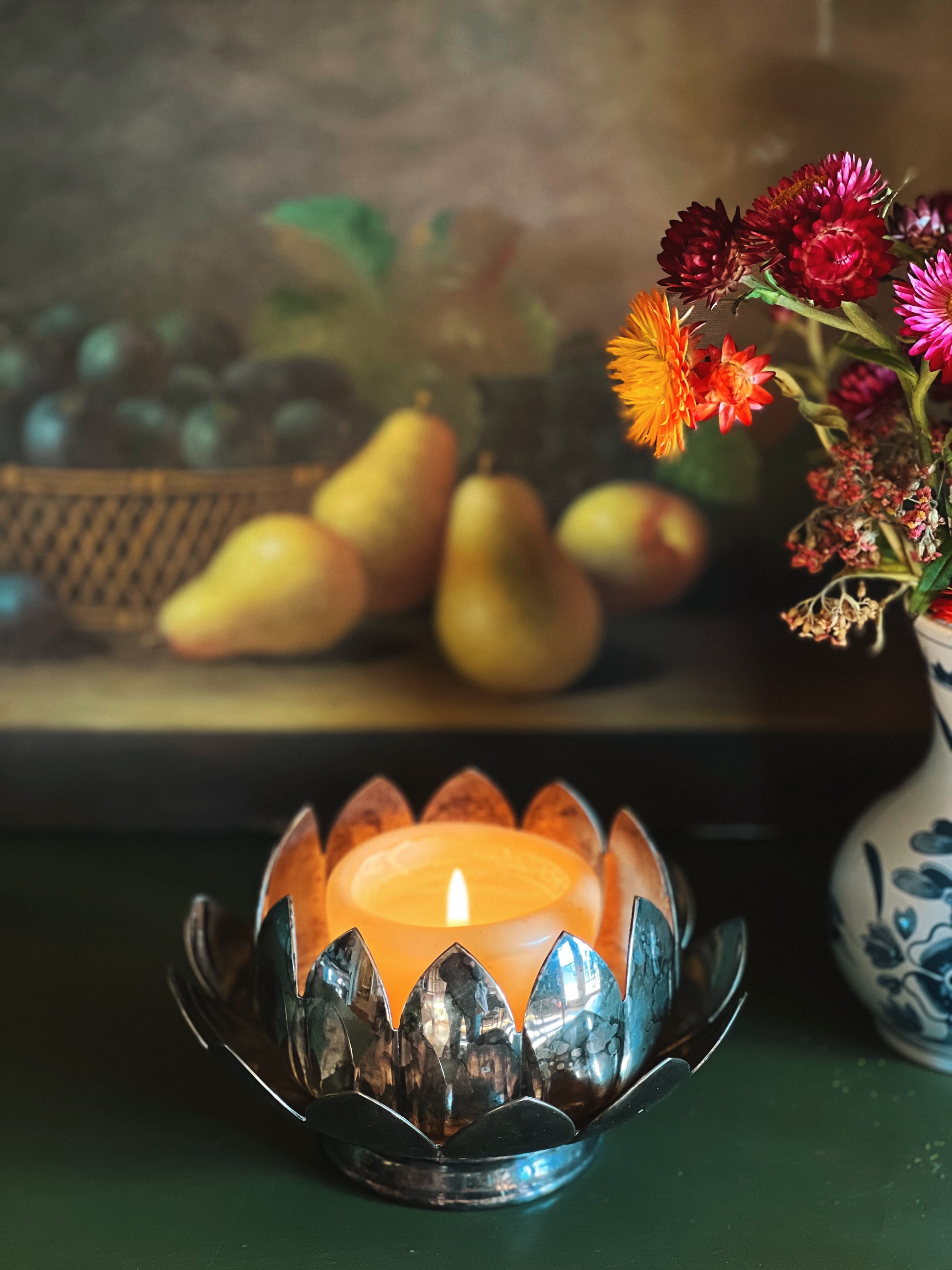 Decorative candle holder with a lit candle, pears, and flowers in the background.