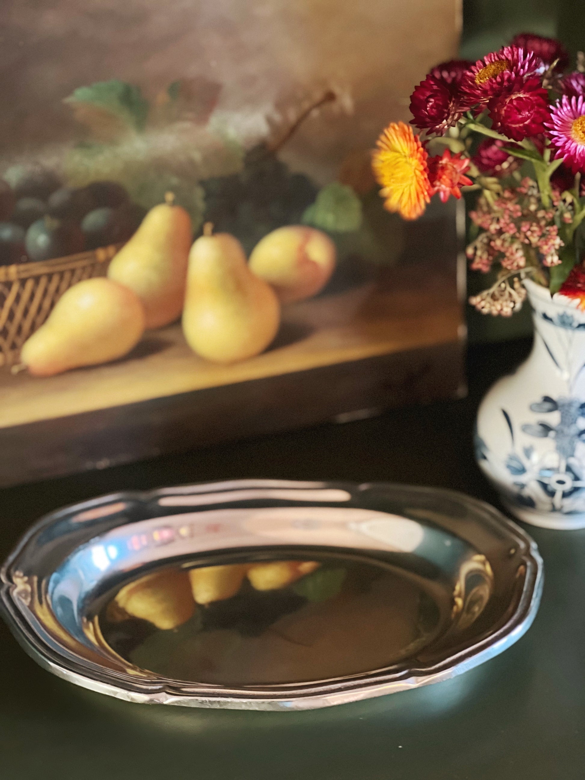 Silver platter on a table with a still life painting and flowers in the background