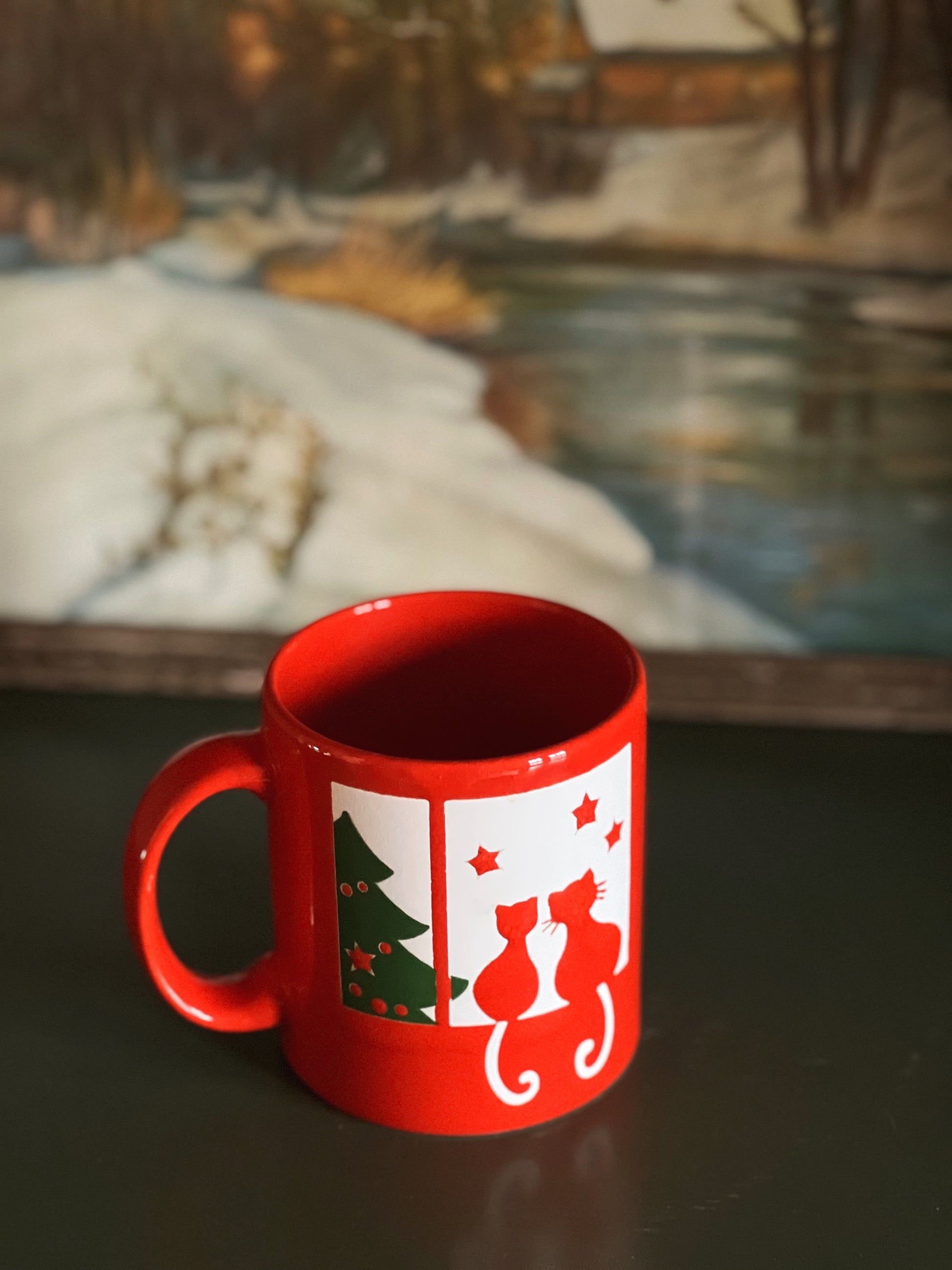 Red mug with a Christmas design featuring two cats and a tree on a dark surface.