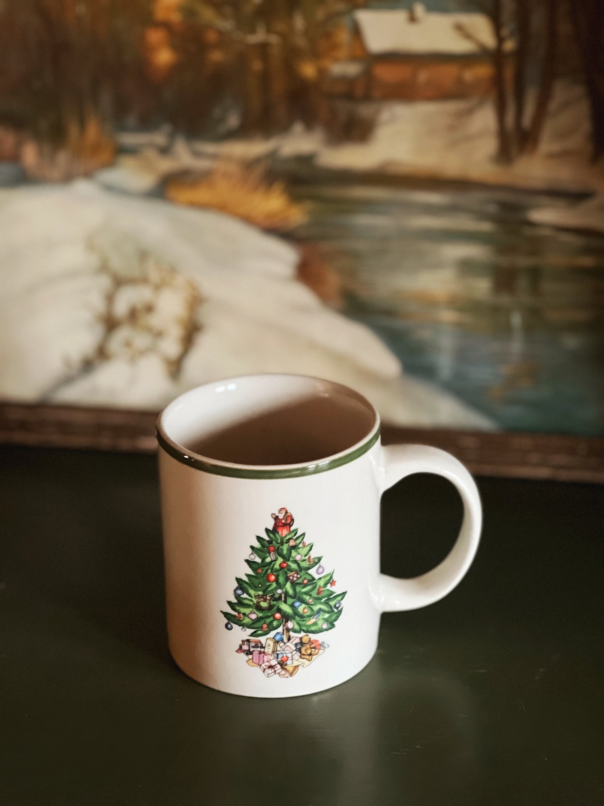 Mug with Christmas tree design on a dark surface, blurred painting in the background