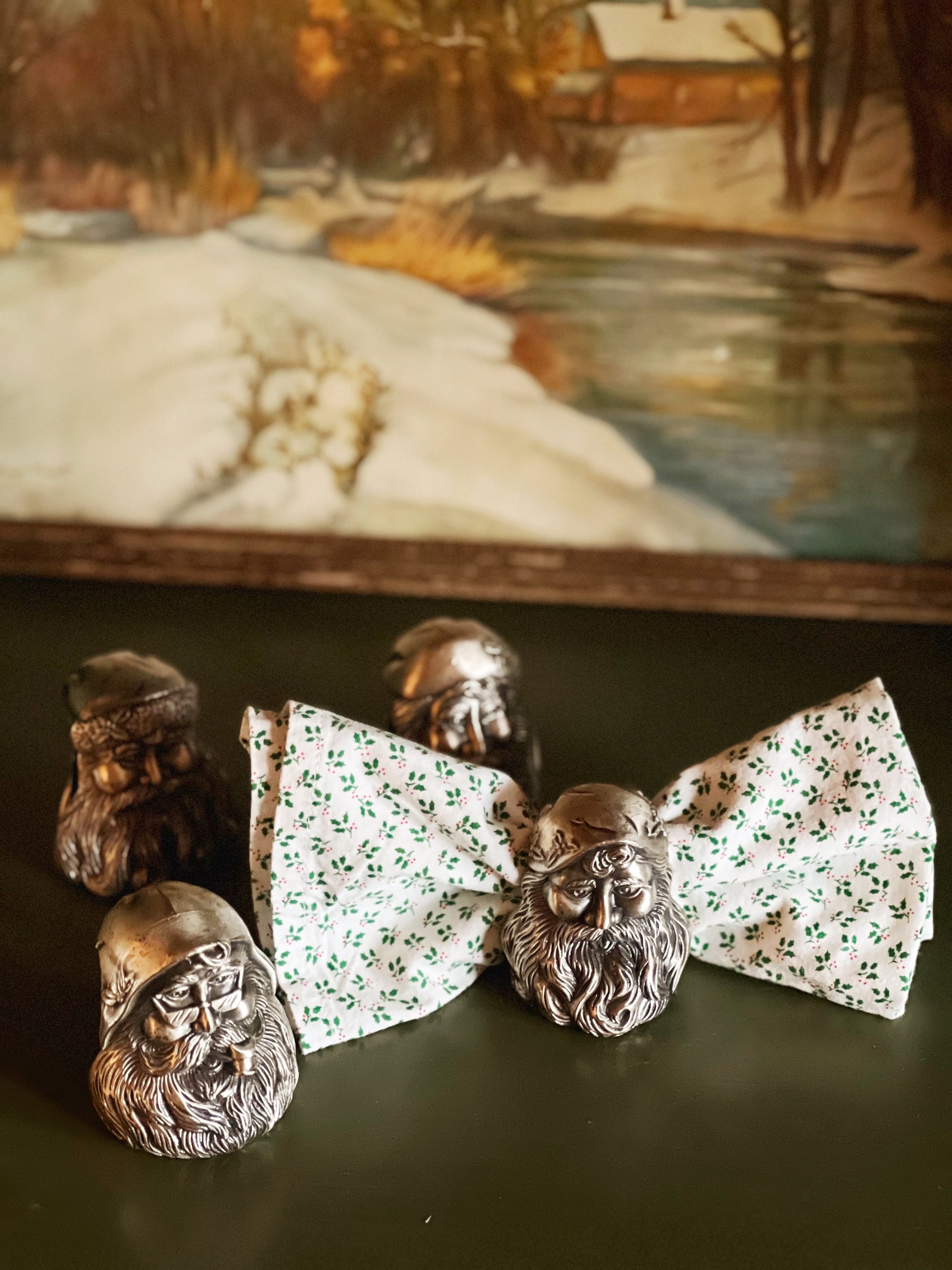 Silver bearded figures with floral-patterned bow ties on a dark surface, with a painting of a snowy landscape in the background.