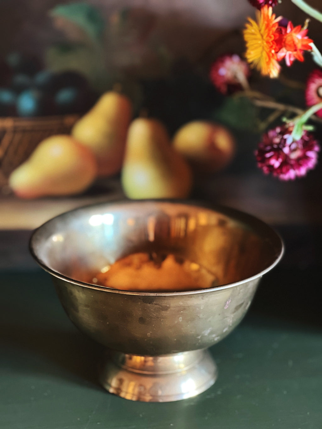 Silver bowl on a dark surface with a blurred background of pears and flowers.