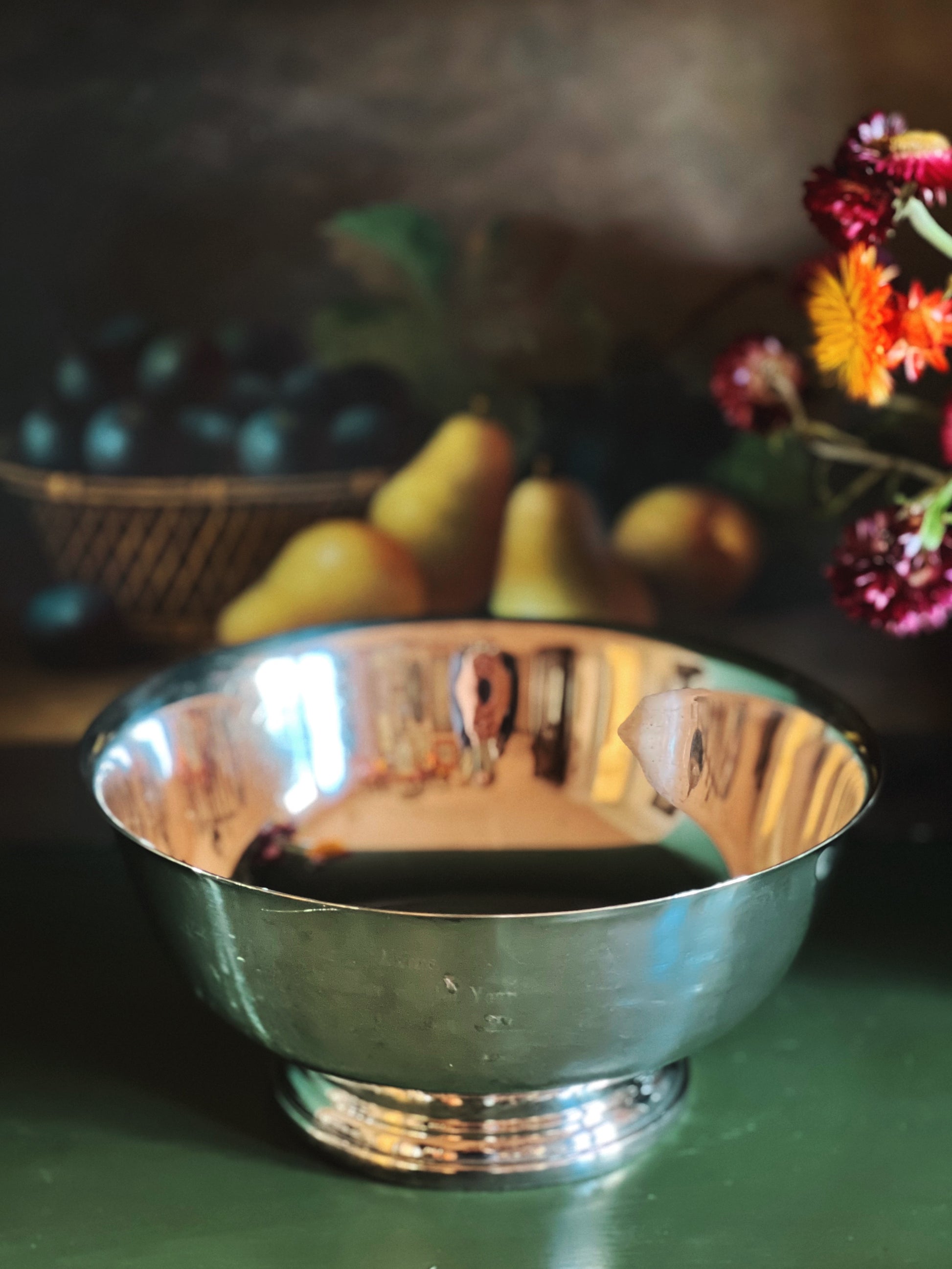 Silver bowl on a table with fruits and flowers in the background