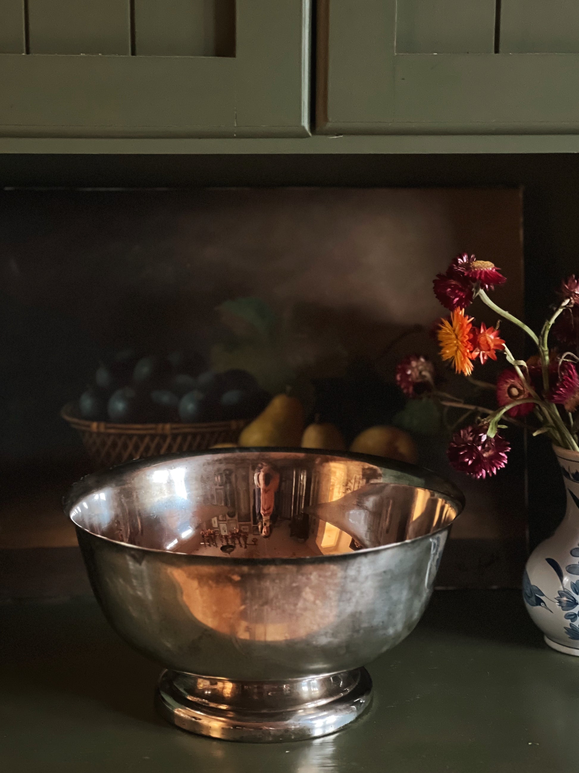 Silver bowl on a wooden surface with a vase of flowers and fruit in the background.