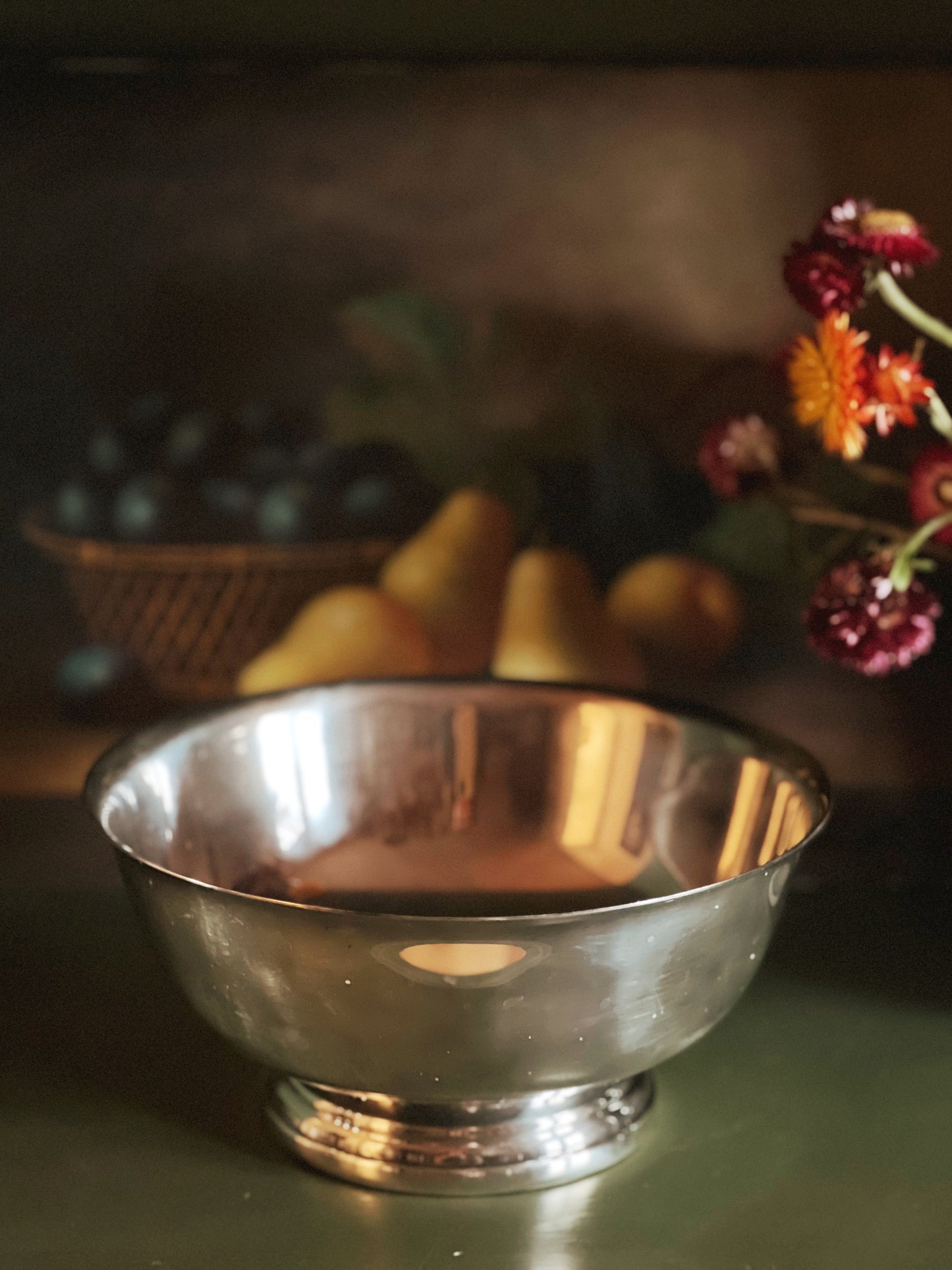 Silver bowl on a table with fruits and flowers in the background