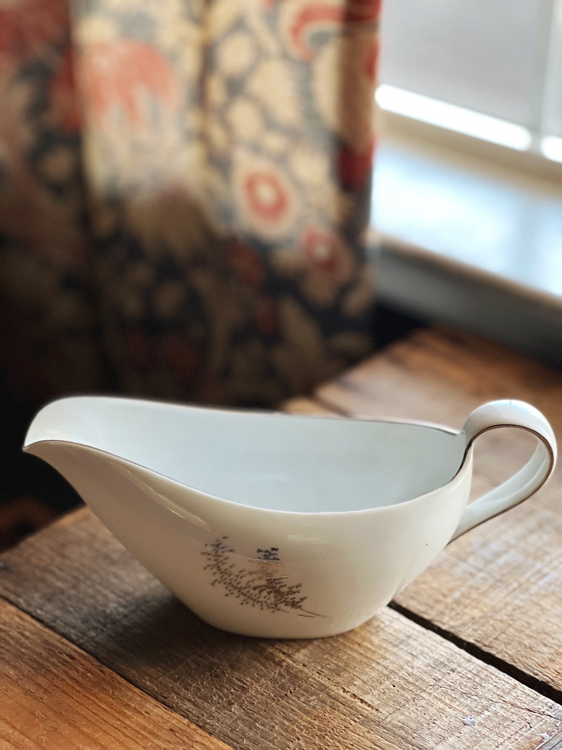 White ceramic gravy boat on a wooden surface with a patterned curtain in the background