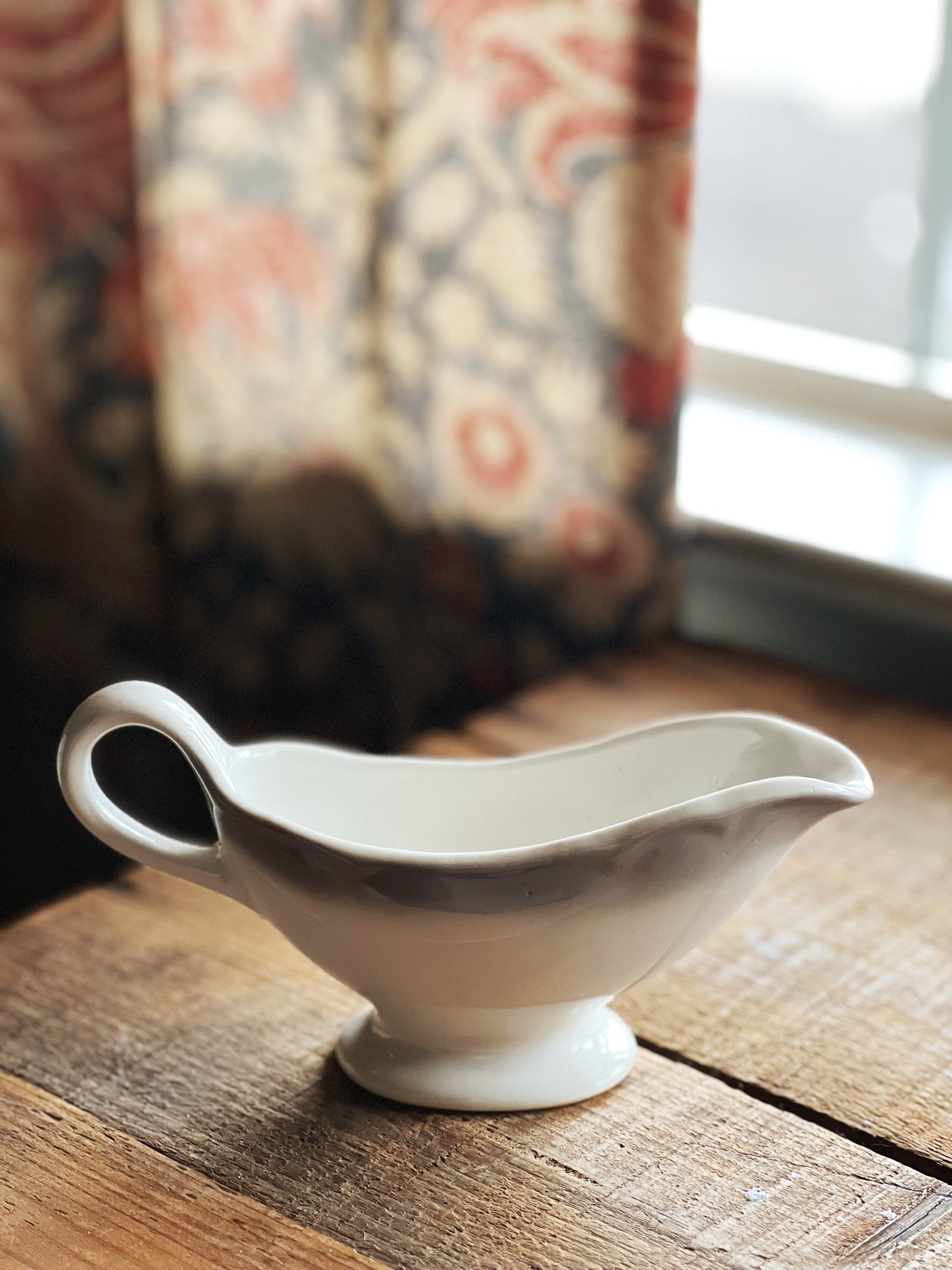 White ceramic gravy boat on a wooden surface with a patterned curtain in the background