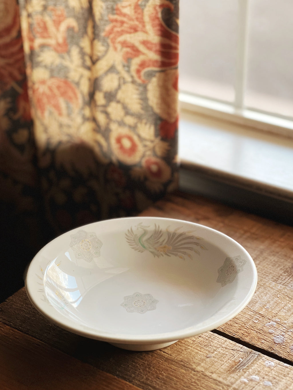 White ceramic bowl with floral pattern on a wooden surface near a window with floral curtains.