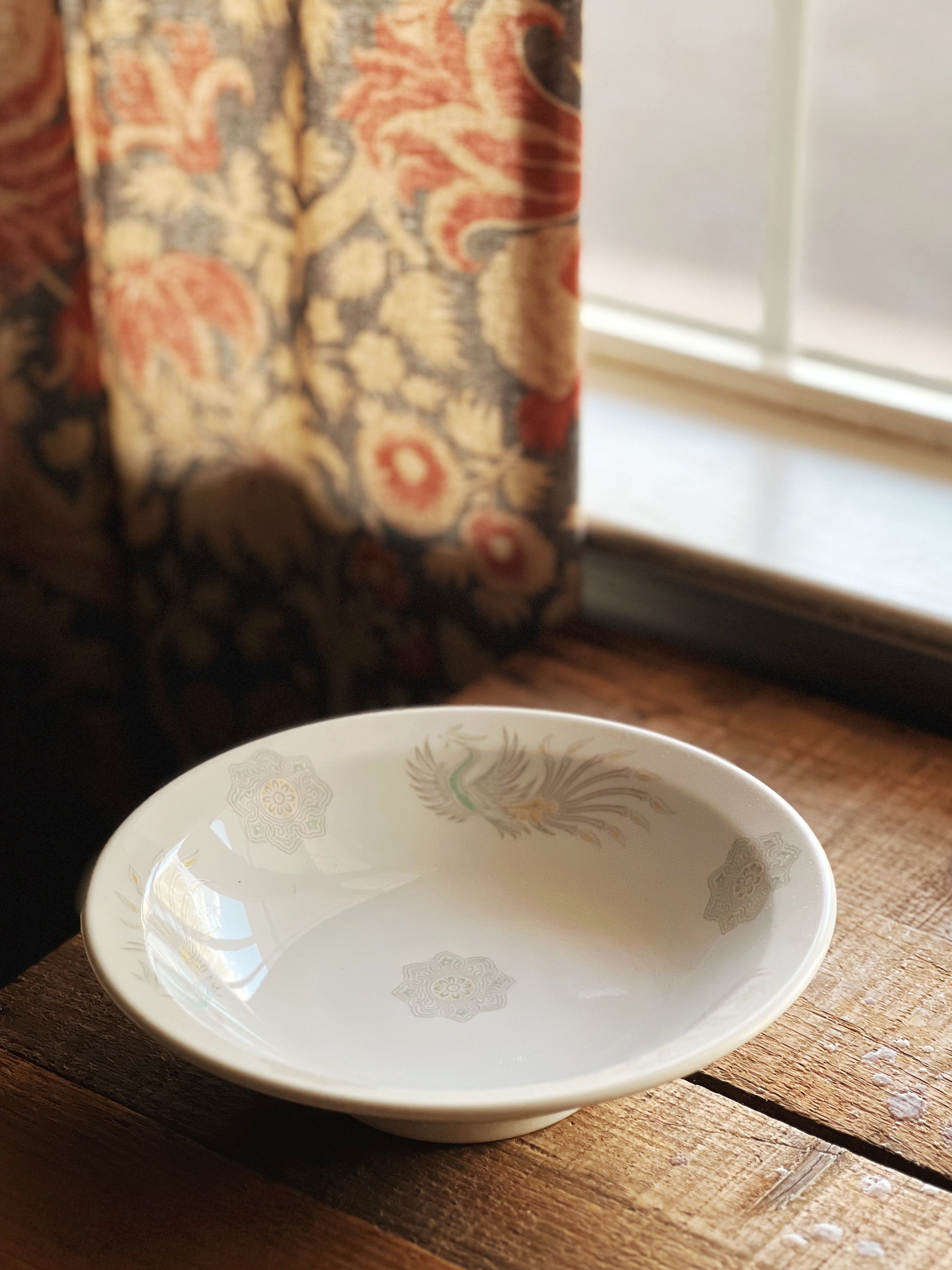 White ceramic bowl with floral pattern on a wooden surface near a window with floral curtains.