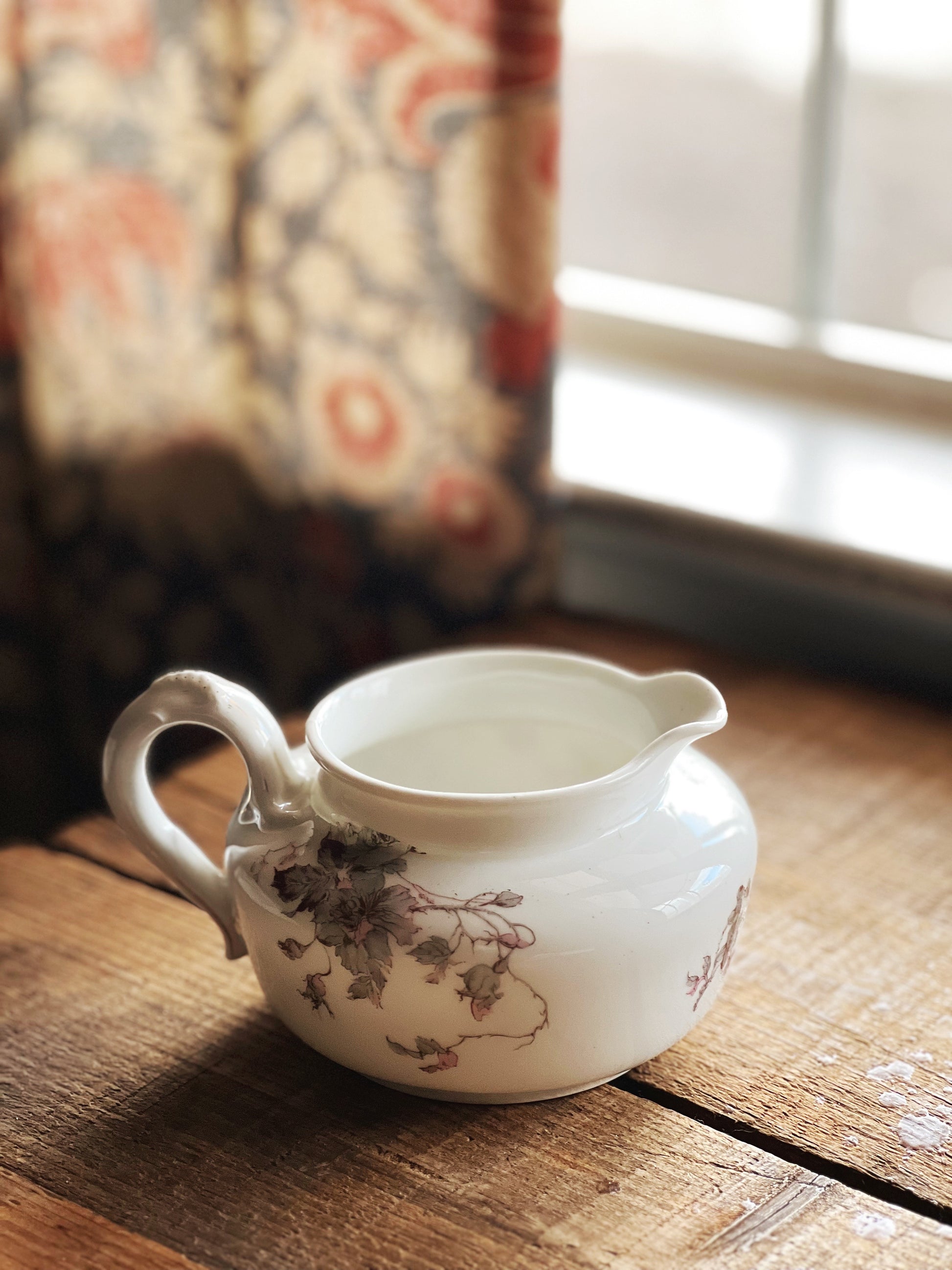 White ceramic pitcher with floral design on a wooden surface near a window with patterned curtains.