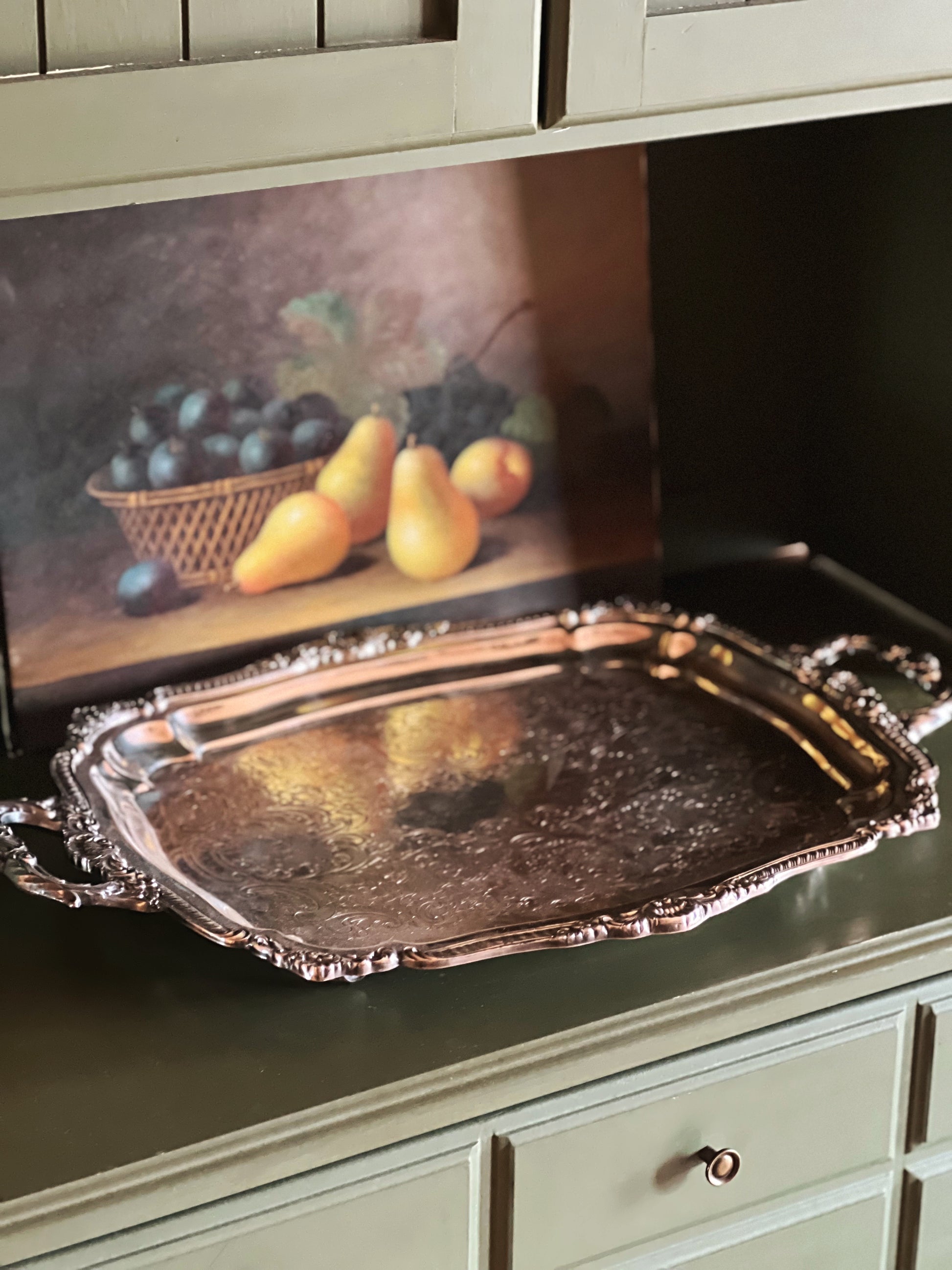 Decorative silver tray on a green cabinet with a painting of fruit in the background